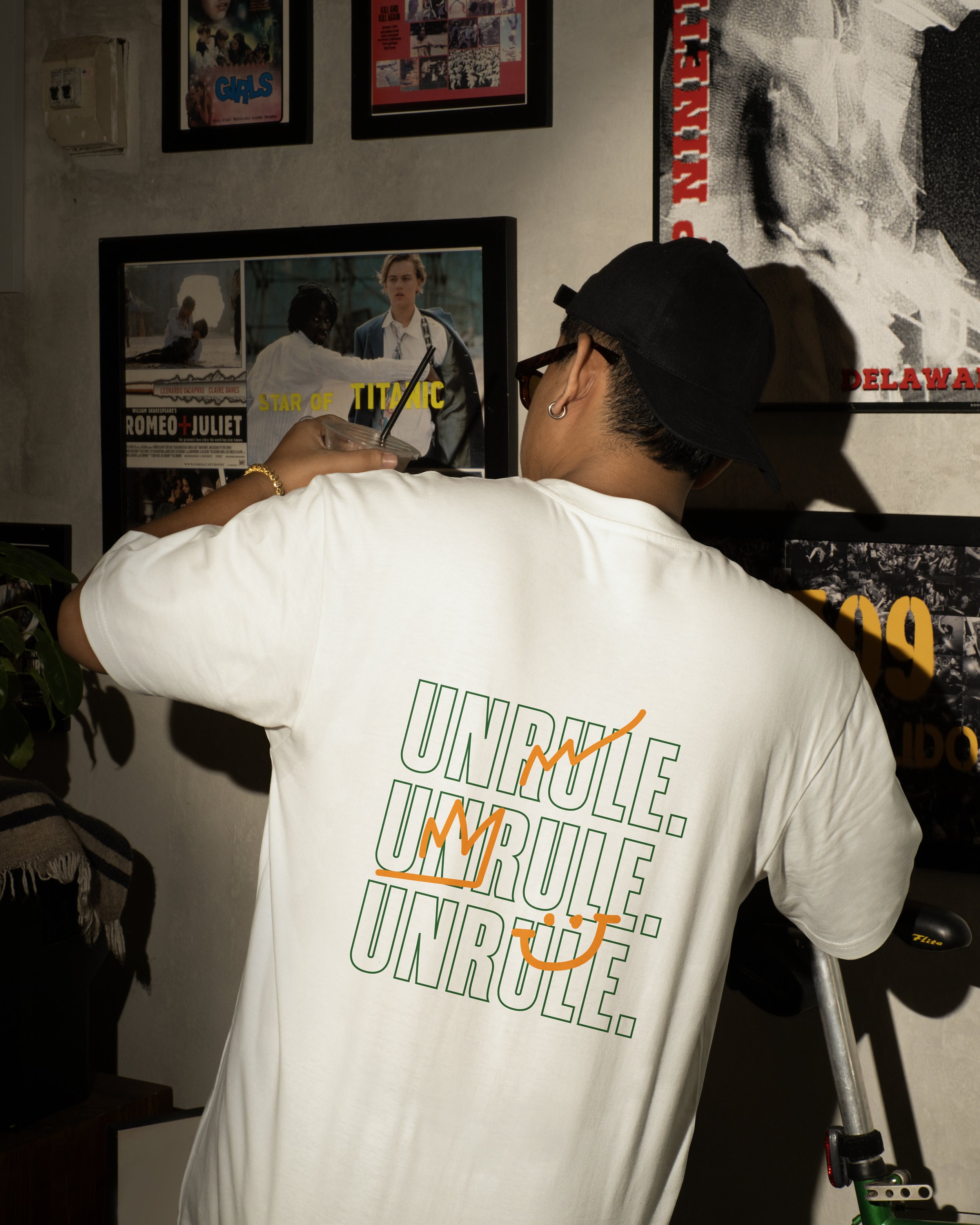 Person wearing a white t-shirt with the text 'UNRULE. UNRULE. UNRULE.' while standing in front of a wall decorated with posters, including a framed 'Star of Titanic' poster and others for movies and bands.
