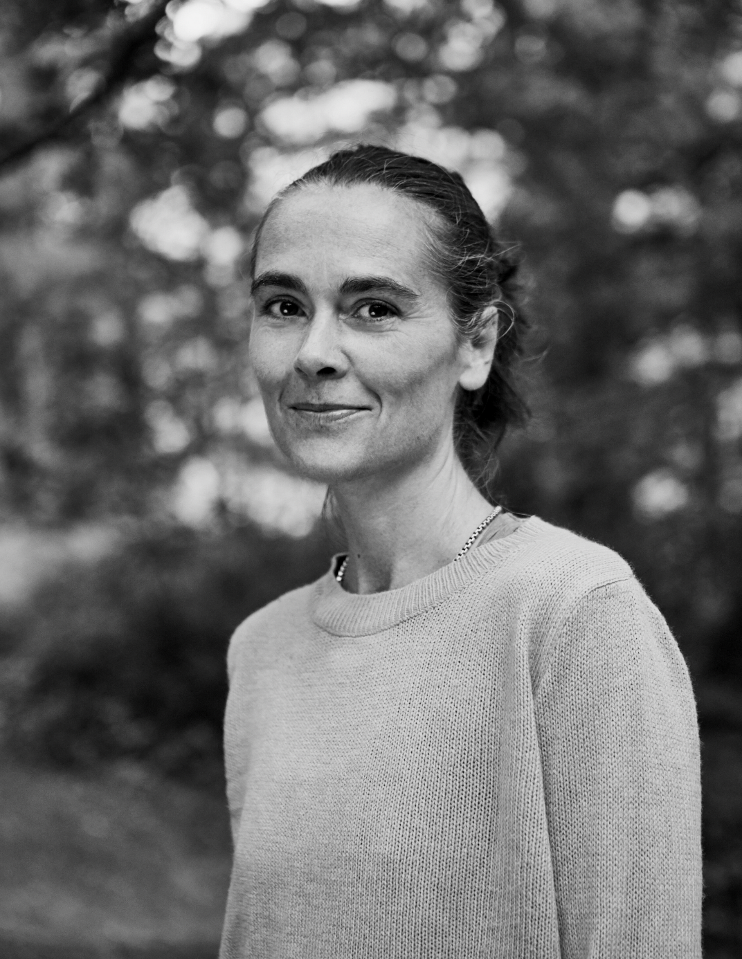 Black and white portrait of a woman with dark hair pulled back, wearing a light-colored sweater and necklace, standing outdoors with blurred trees in the background.