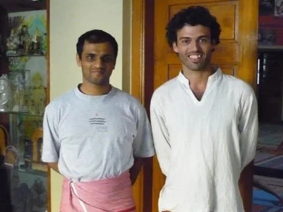 Two men standing indoors, one wearing a gray t-shirt and pink apron, the other in a white shirt, smiling.