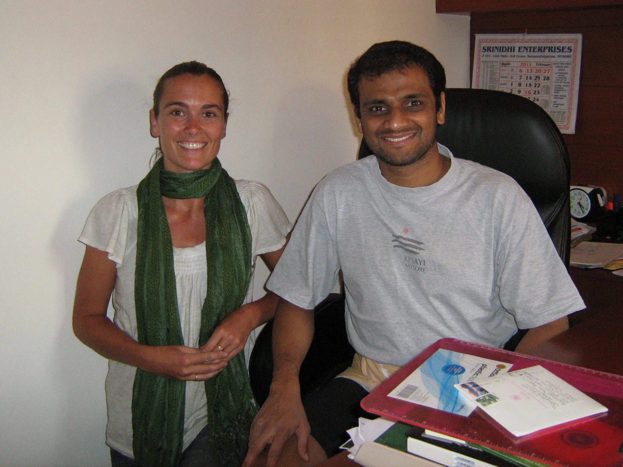 A woman and a man smiling and posing for a photo in an office setting, with a desk and calendar in the background.