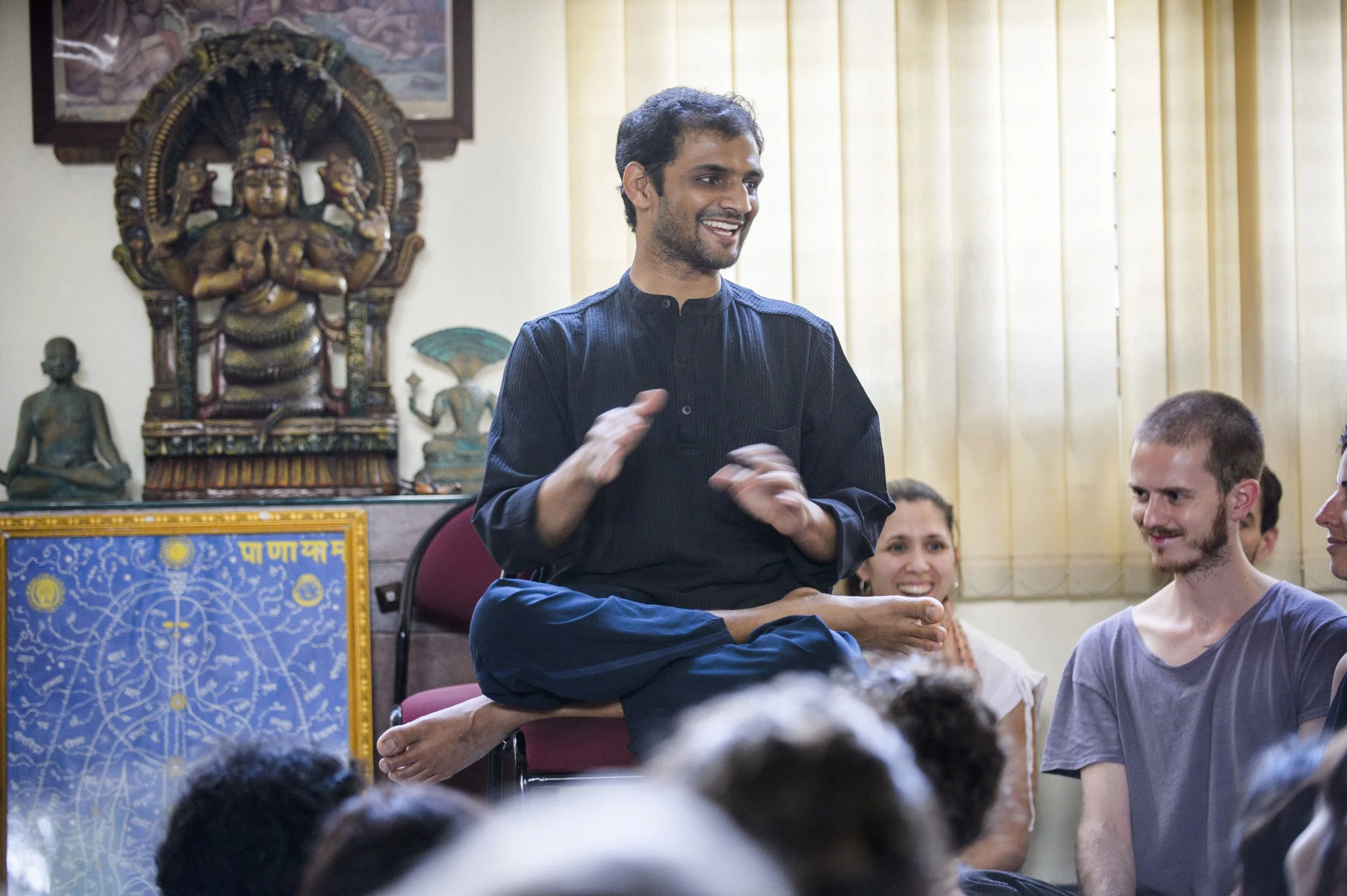A man sitting cross-legged on a chair, smiling and gesturing with his hands, while three people sit in front of him, smiling and listening. In the background, there are religious statues and artwork, including a large Hindu deity sculpture and a colorful painting with Tibetan script.