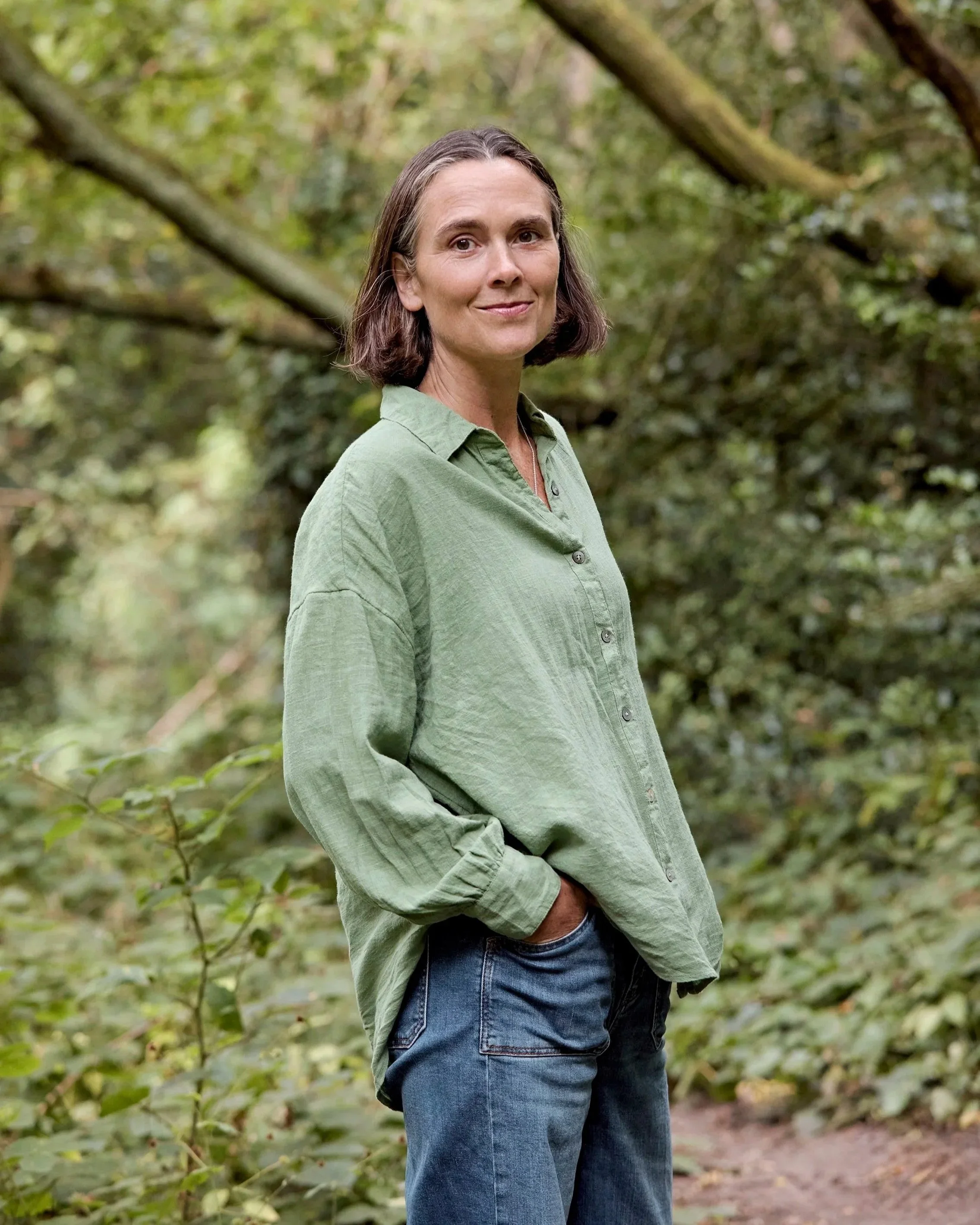 A woman with shoulder-length brown hair, wearing a loose-fitting green shirt and blue jeans, standing outdoors in a wooded area with green leaves and branches in the background.