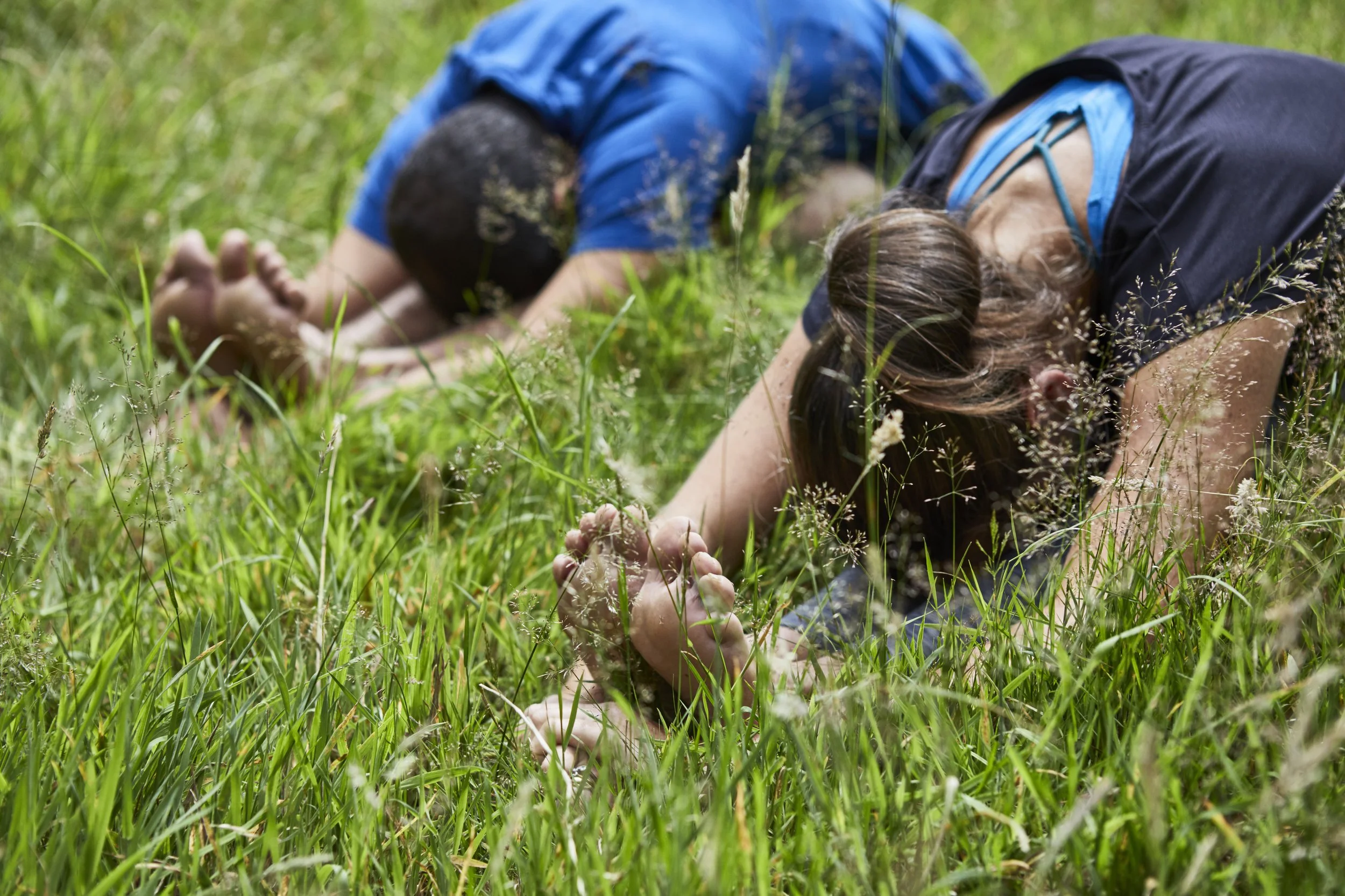 Two people practicing yoga outdoors in a grassy field, both in child's pose with arms extended forward and heads down.