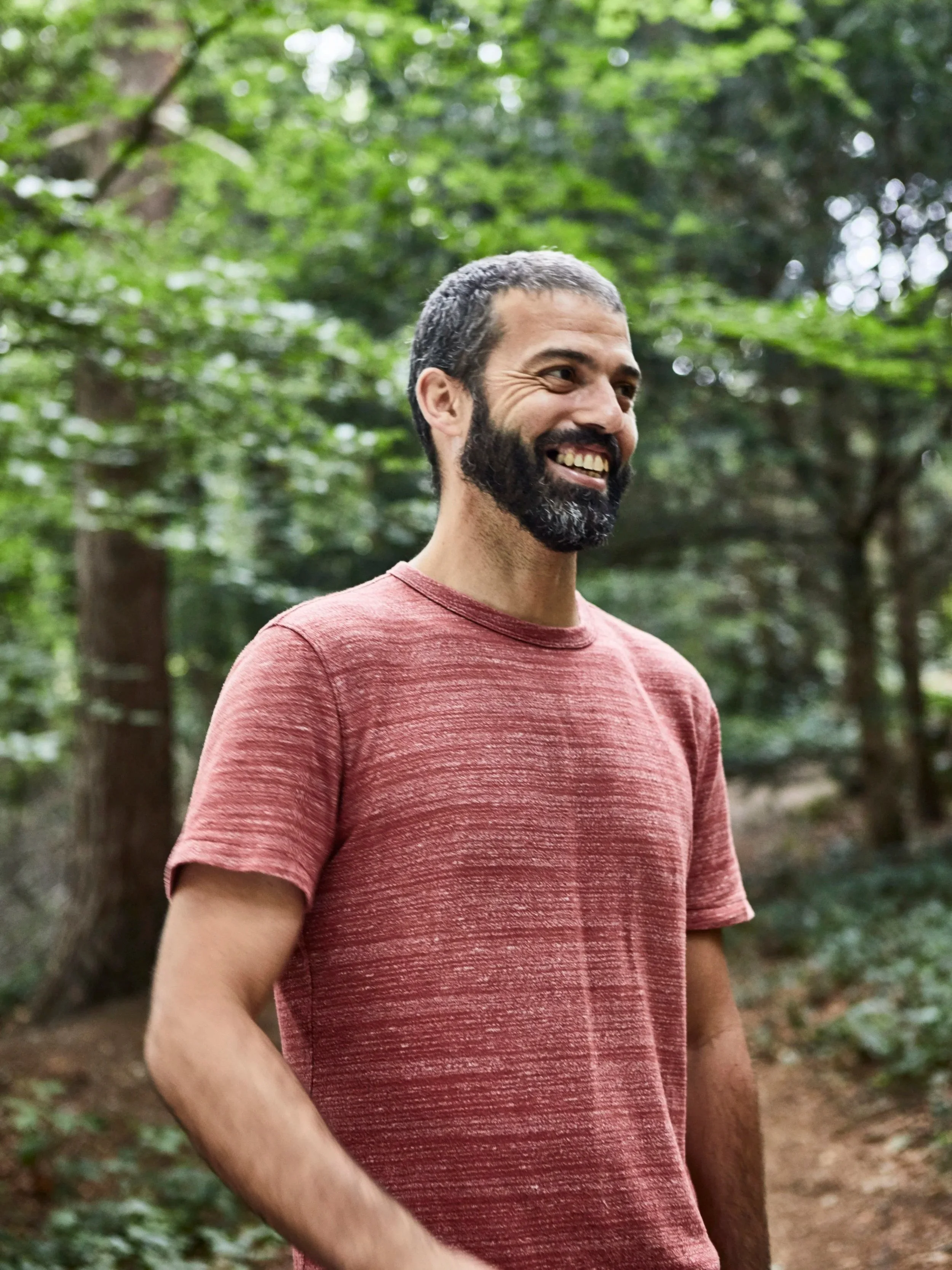 A man with short dark hair, wearing a red T-shirt, smiling in a wooded area with green trees in the background.