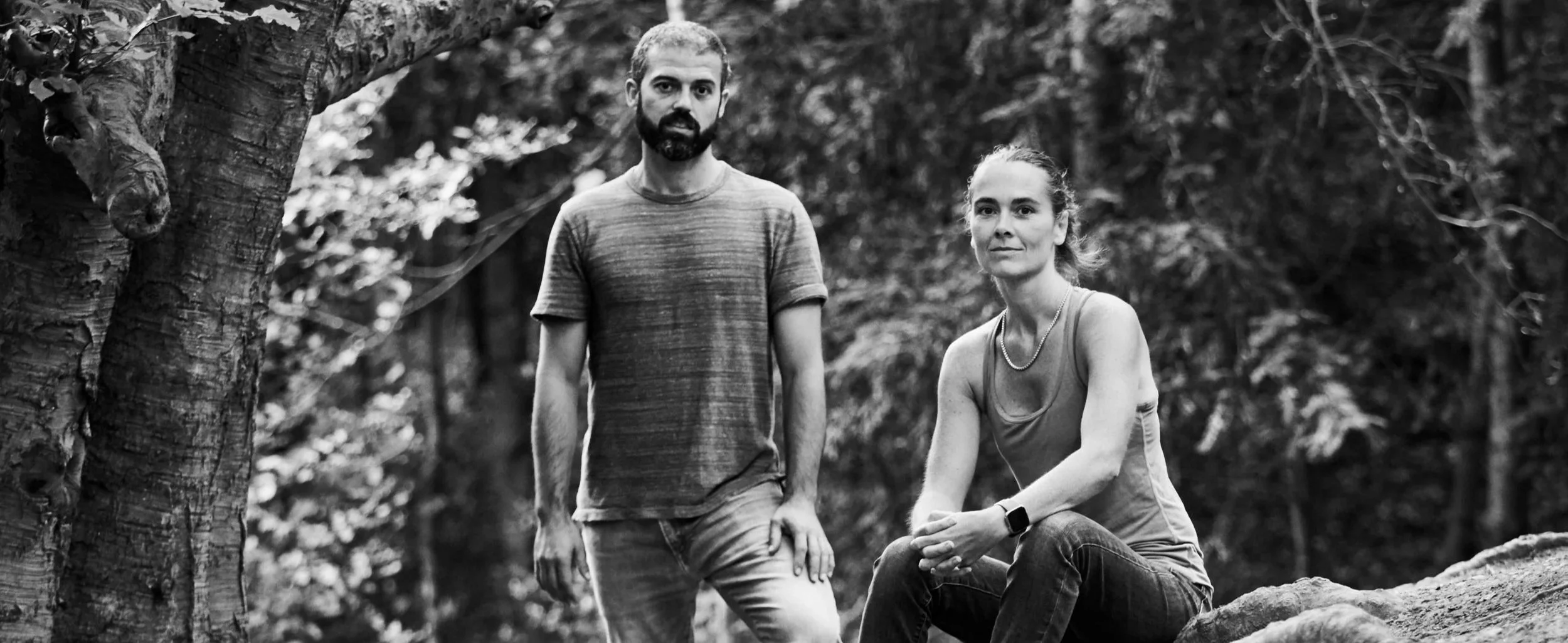 A man with a beard and a woman with short hair in a wooded area, standing and sitting on a log, both looking at the camera in black and white.