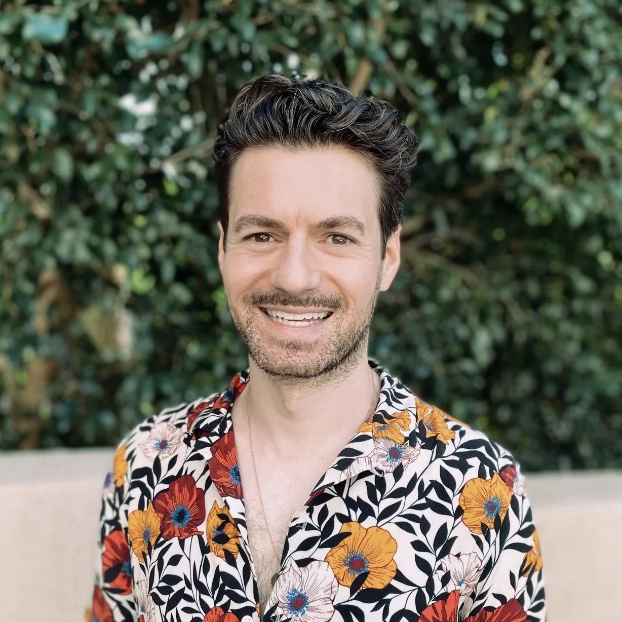 A smiling man with dark hair and stubble, wearing a colorful floral shirt, standing outdoors in front of green foliage.