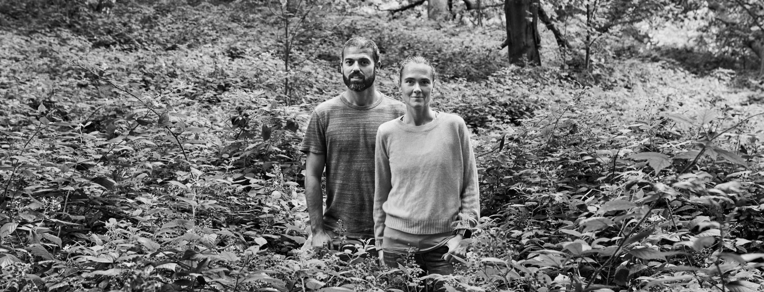A black and white photo of a man and woman standing in a dense forest or bush, facing the camera.