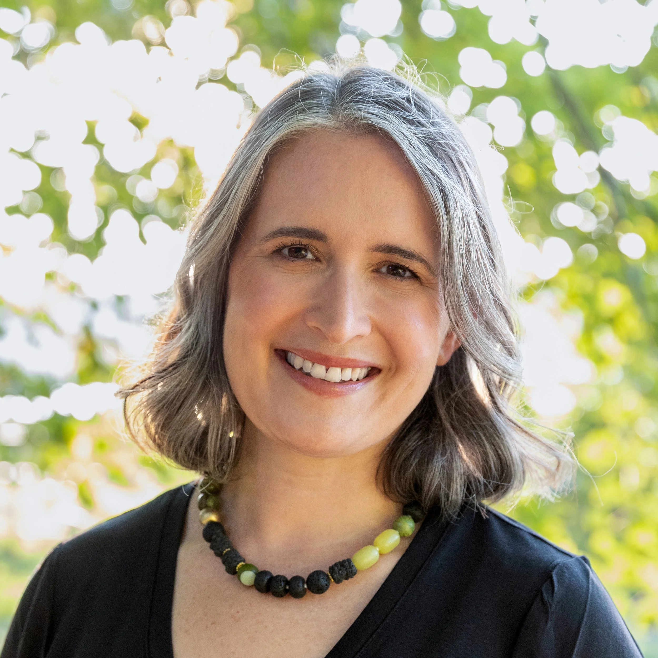 A woman with shoulder-length gray hair smiling outdoors, wearing a black top and a necklace with beads, with green foliage and sunlight in the background.