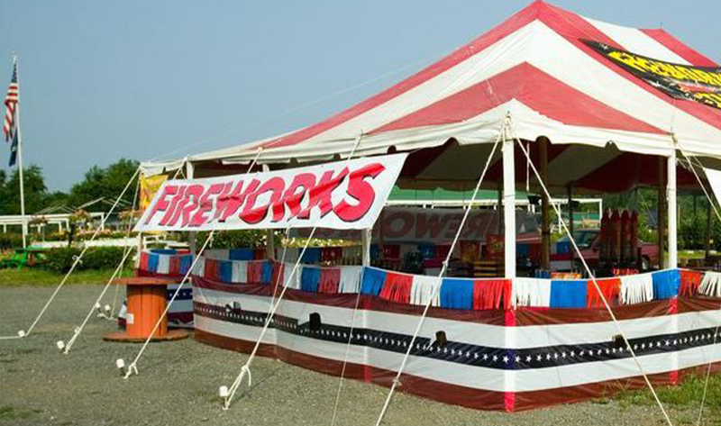 A red and white striped tent with a sign that says 'FIREWORKS' at an outdoor event, with fireworks display banners on top.