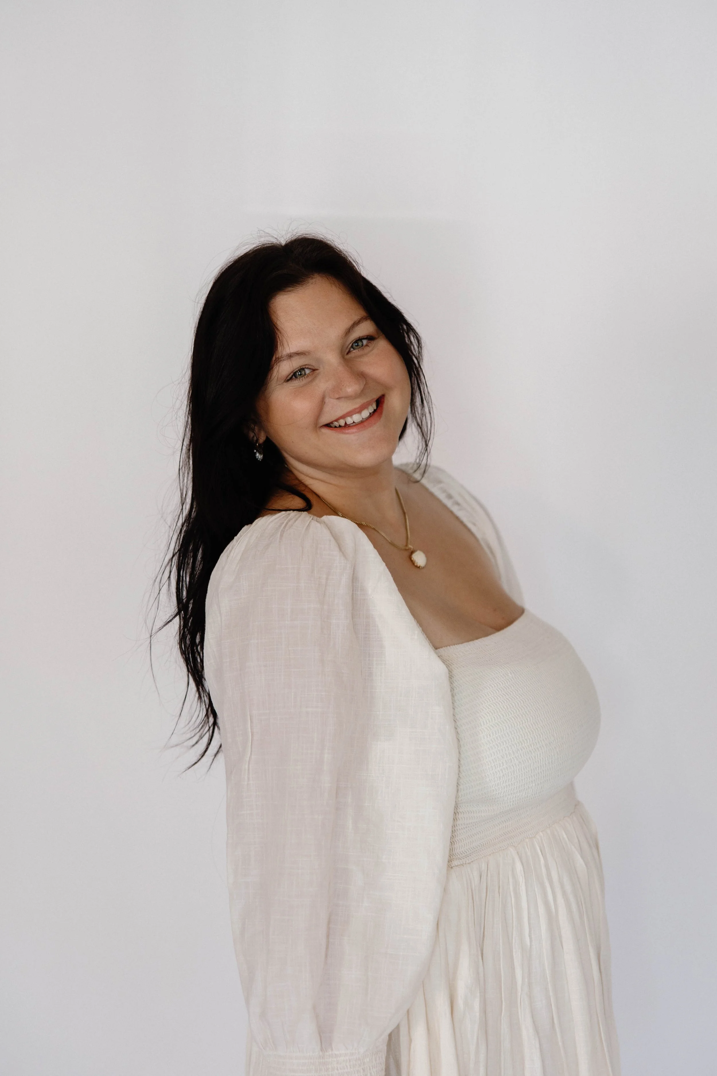 A woman with long dark hair smiling, wearing a cream-colored dress and a necklace, standing against a plain white background.