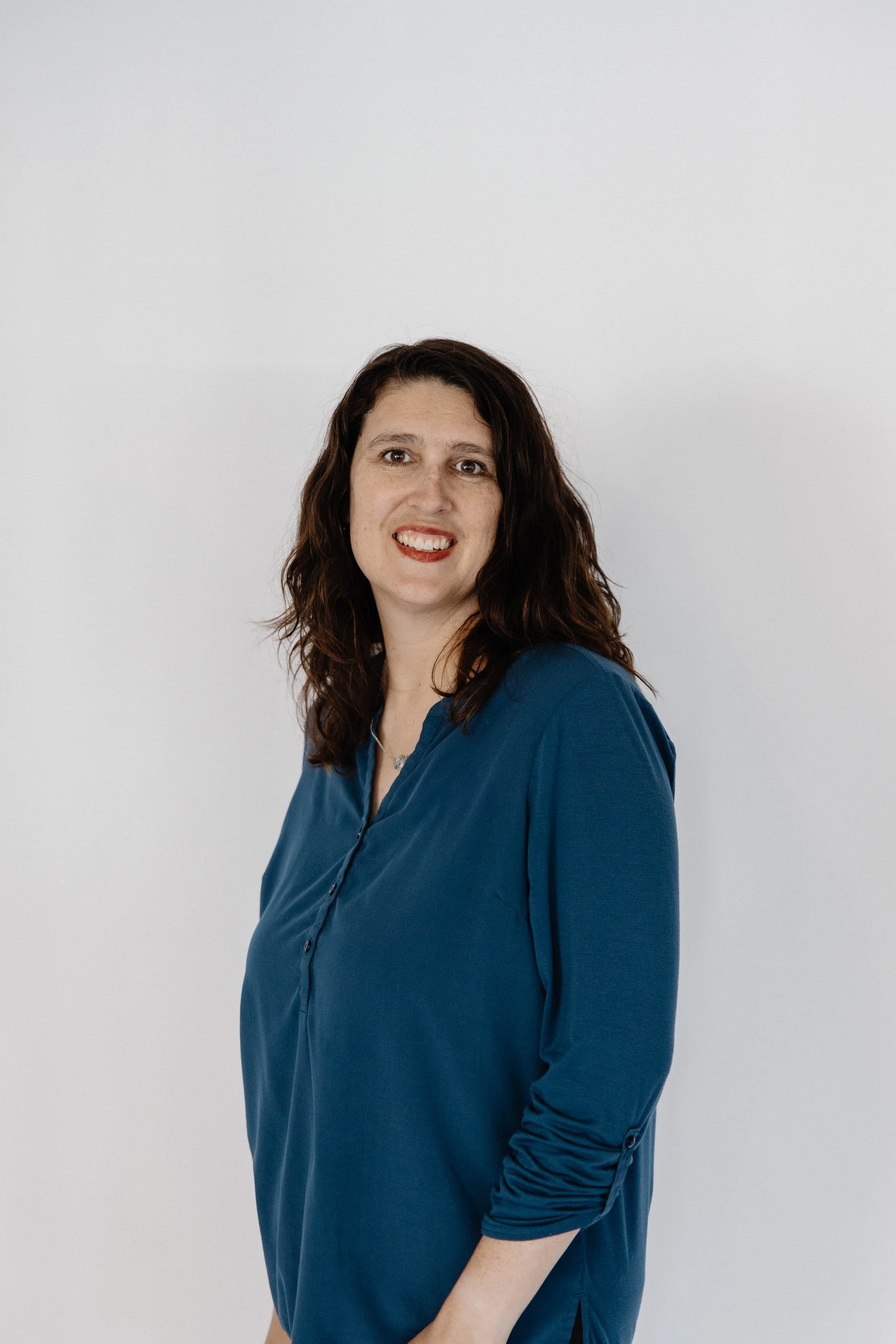 A woman with brown, wavy hair wearing a blue shirt, standing against a plain white background.