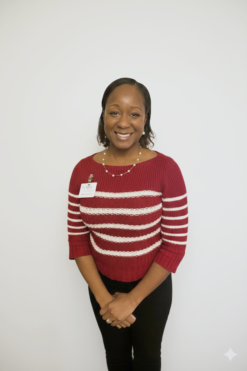 A woman standing against a plain white wall, smiling, wearing a red and white striped sweater, black pants, a pearl necklace, and a name badge.