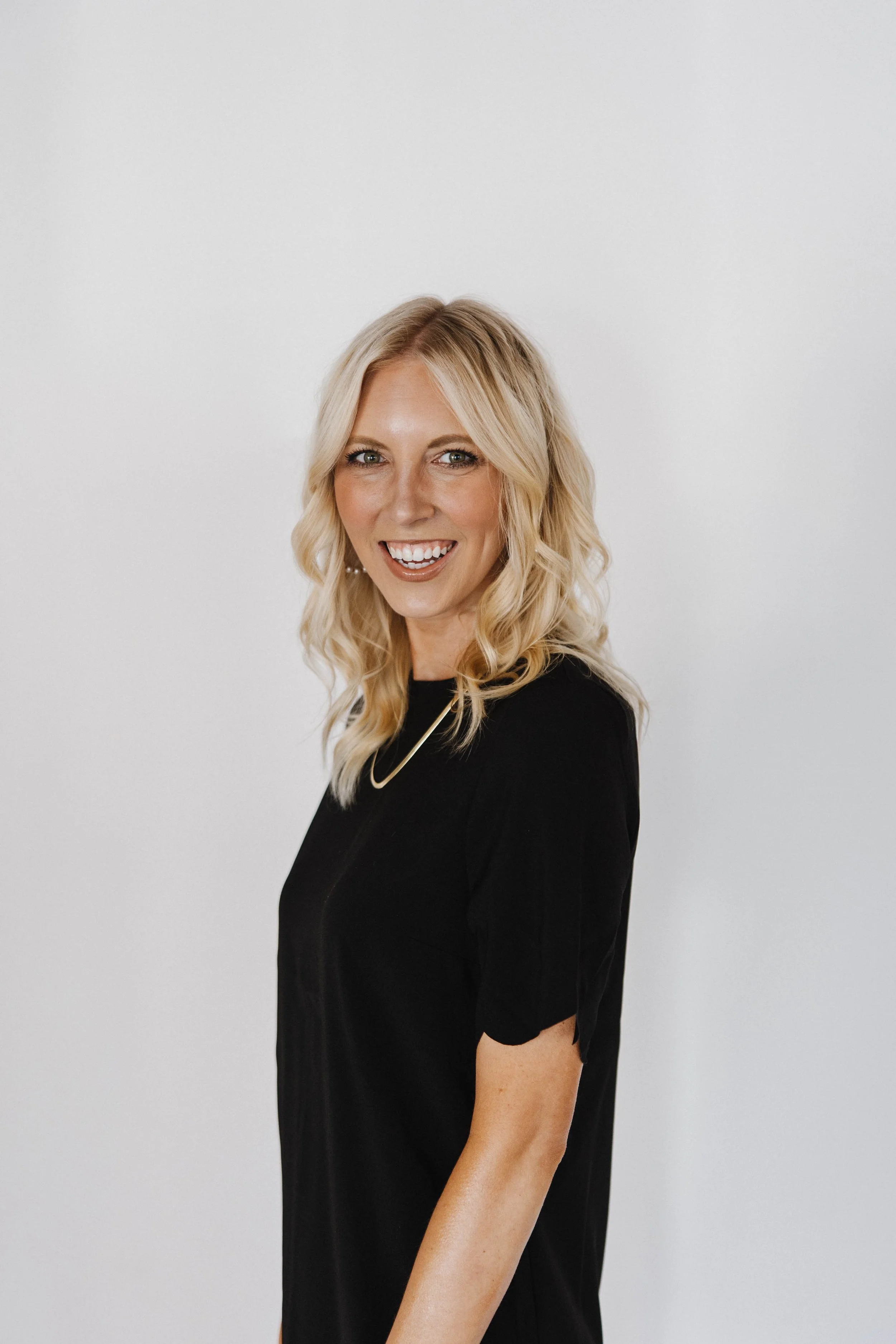 A woman with blonde wavy hair wearing a black top, smiling and looking at the camera against a plain white background.