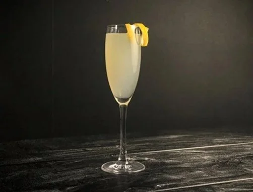 A champagne glass with SideBar® Secret Cove Lime and sparkling wine with a lemon twist garnish, on a dark wooden surface against a black background.