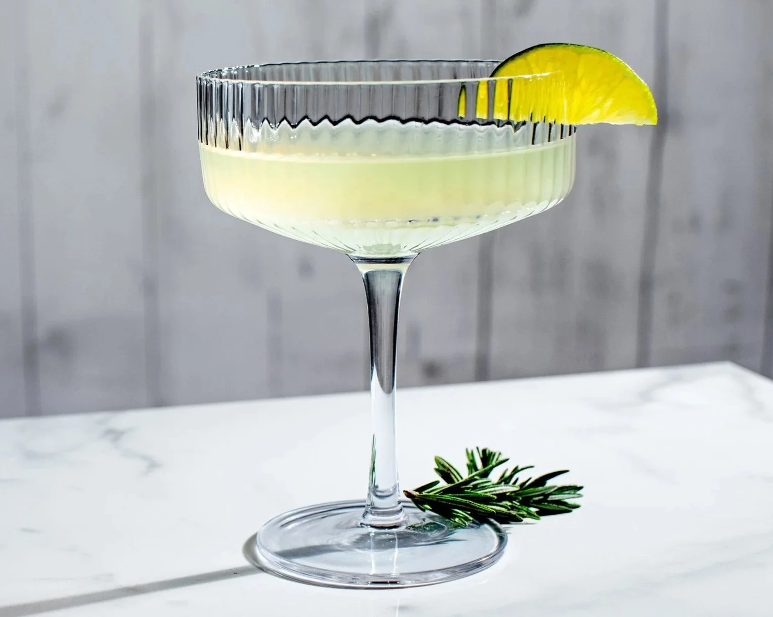 A coupe cocktail glass with a SideBar® Secret Cove Lime garnished with a lemon wedge, resting on a white surface with a sprig of rosemary.