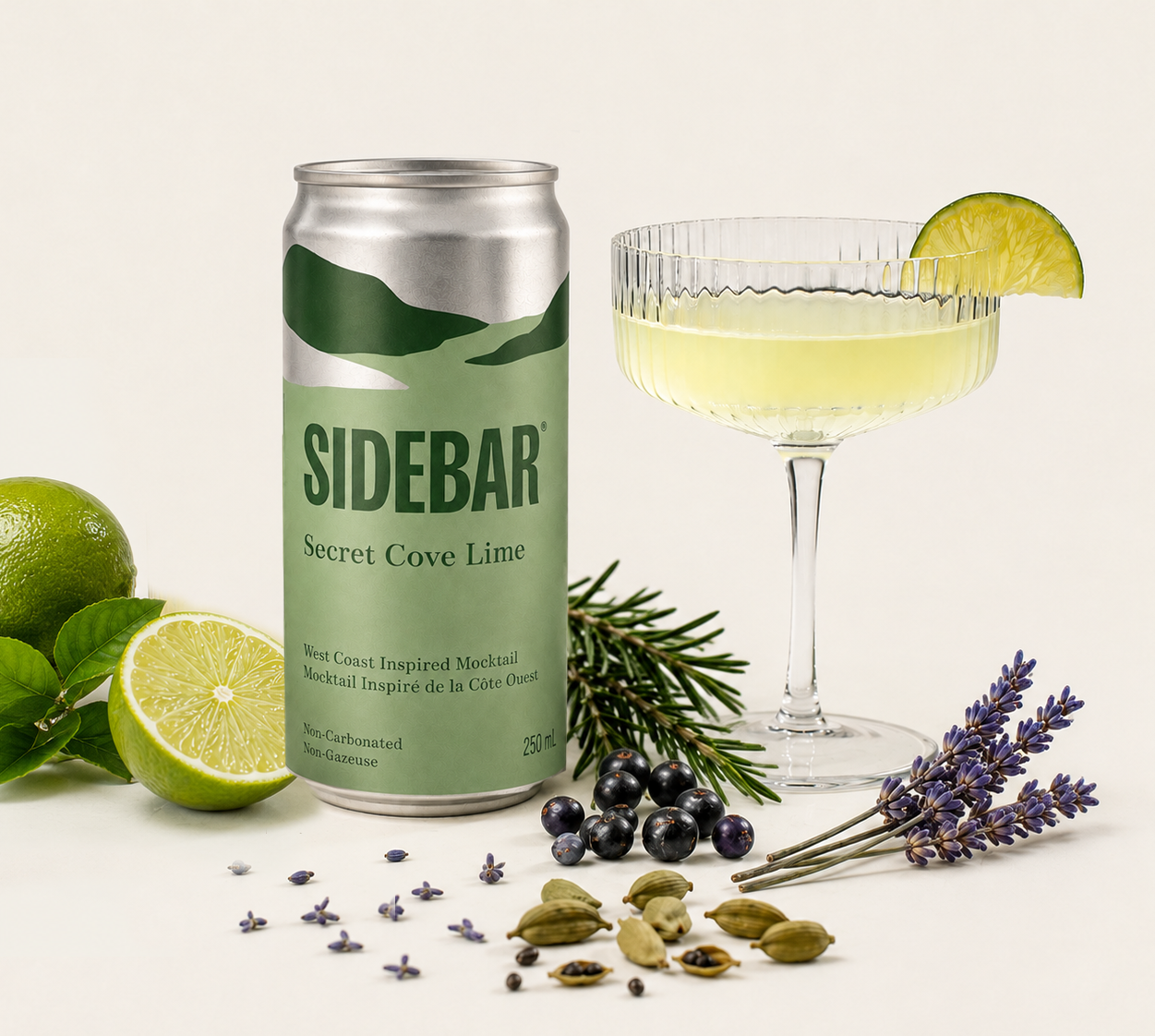 A can of SideBar® Secret Cove Lime mocktail next to a cocktail glass with SideBar® Secret Cove with a lime wedge, surrounded by limes, juniper, lavender sprigs, and cardamon.