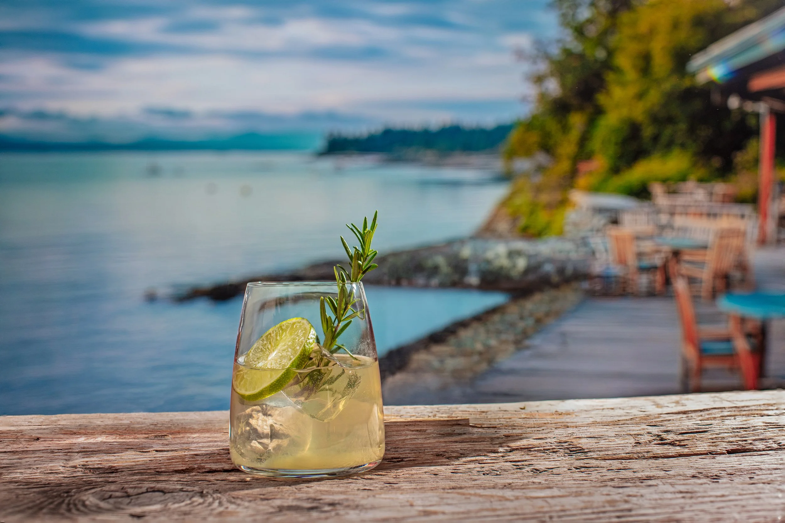 A glass of SideBar® Secret Cove Lime with a lime slice and sprig of rosemary on a wooden surface, overlooking a rocky coastline, trees and boats on the water.