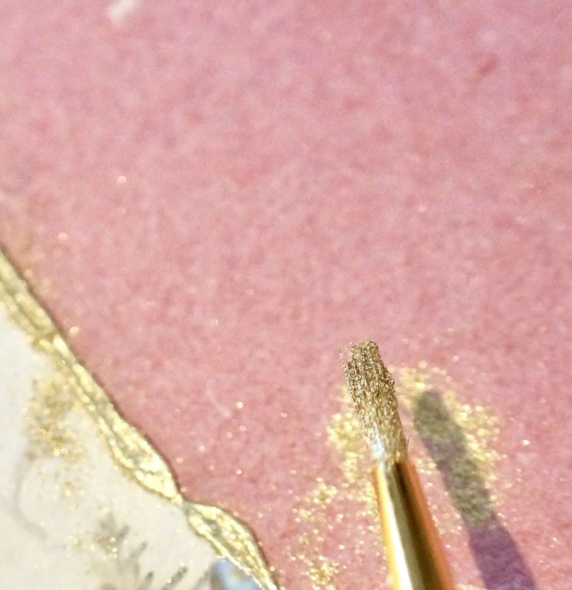 Close-up of a makeup brush applying pink eyeshadow with gold glitter on eyelid, with a gold-edged eyeshadow palette partially visible in the corner.