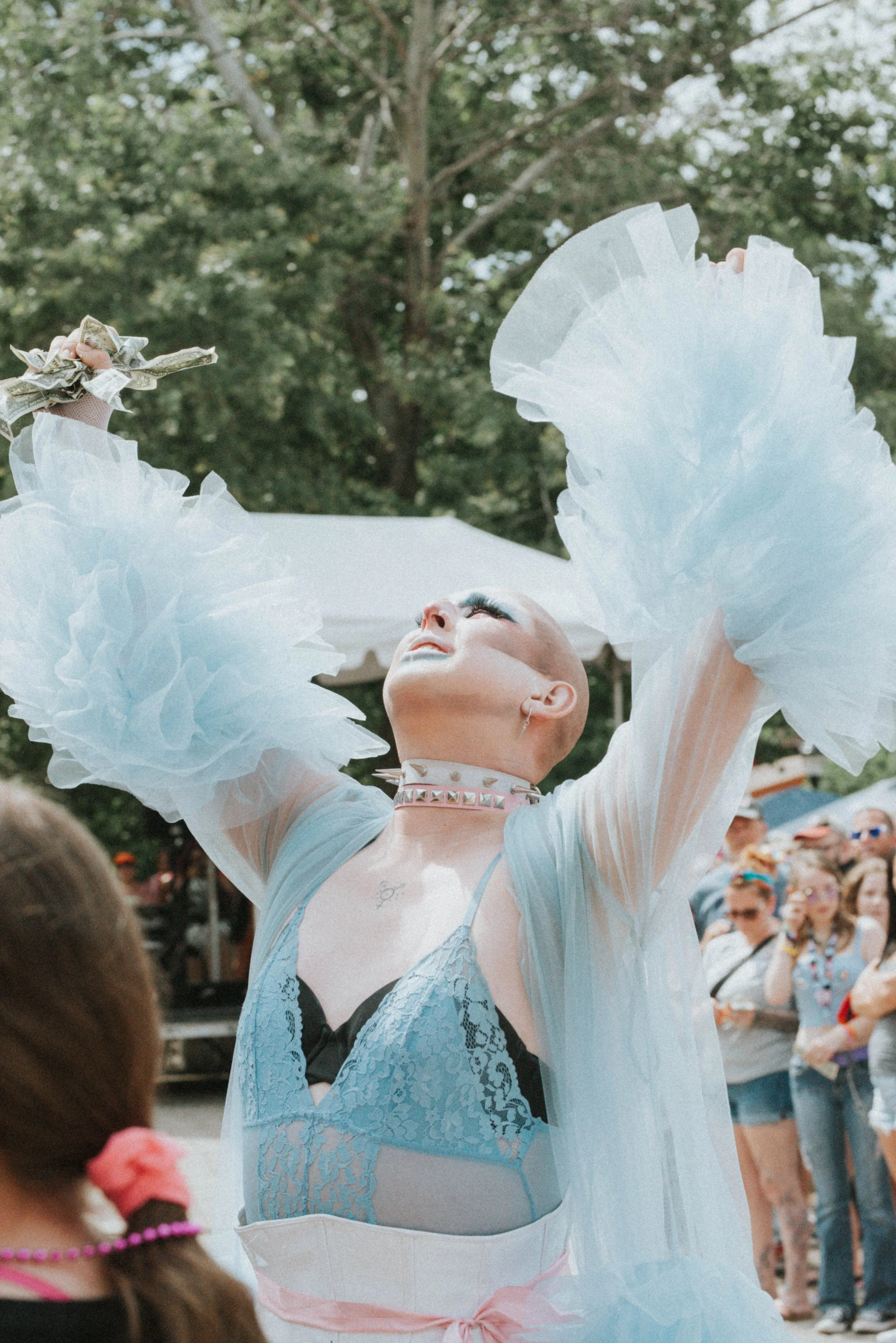 A person with a shaved head wearing a pastel blue and black lace outfit, with large white tulle ruffles on their arms, performing a dance or show outdoors. They are holding money in their hand, with a crowd of onlookers and green trees in the background.