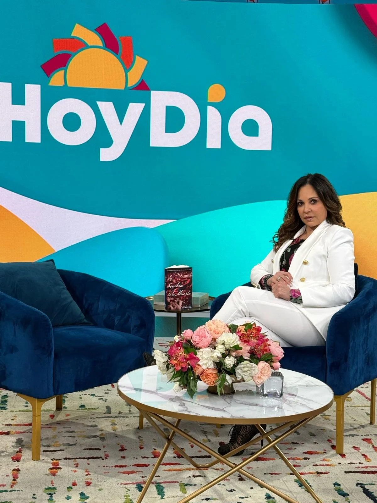 Nenita Foundation founder Krystal Fernandez sits on a blue velvet armchair in front of colorful “Hoy Día” tv set with a sun illustration. She wears a white suit, beside a marble coffee table with flowers and a book.