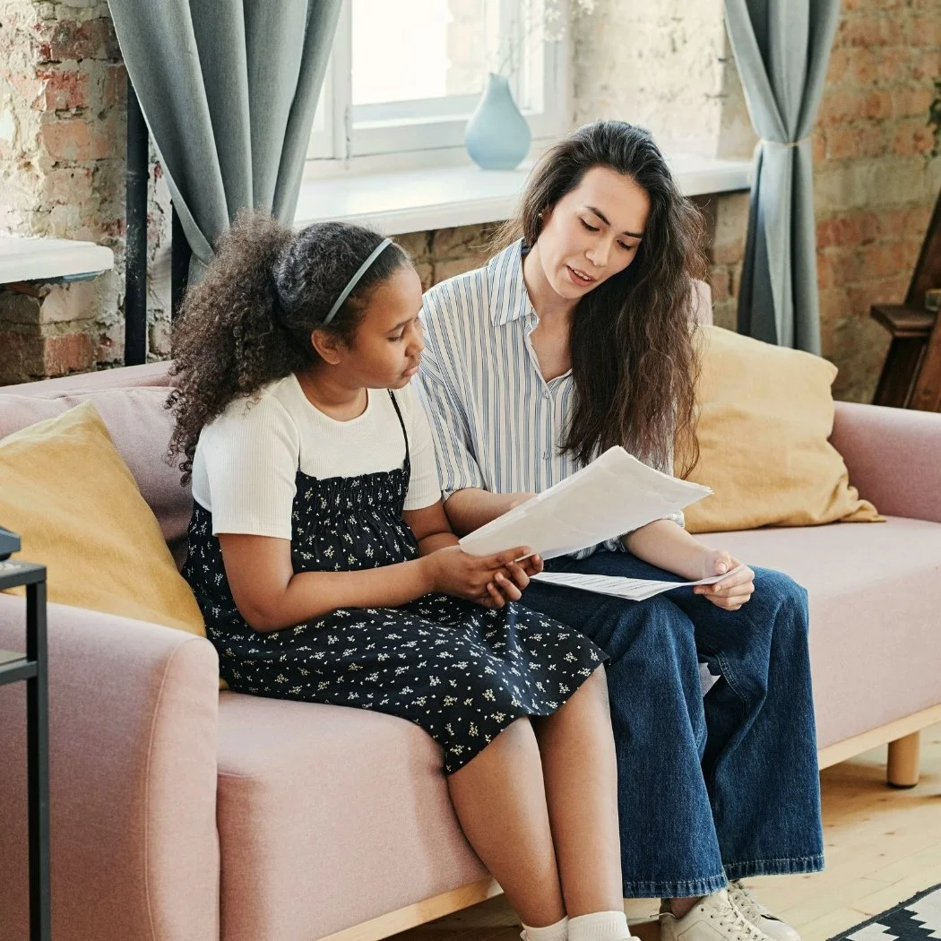 A woman and young girl sitting on a pink sofa reviewing papers together, symbolizing The Nenita Foundation’s focus on mental health support, trauma support, and emotional healing.