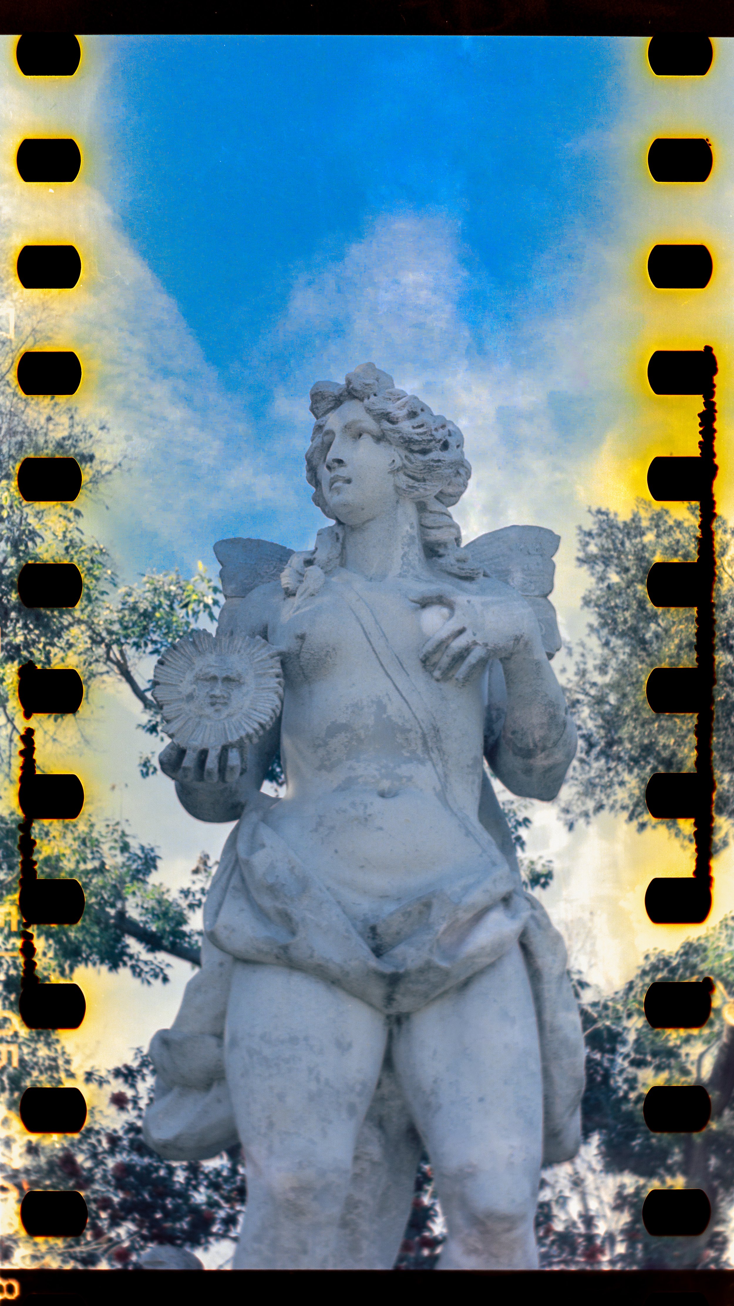 Statue of a winged female figure holding a sun with a face, outdoors with trees and a partly cloudy sky in the background.