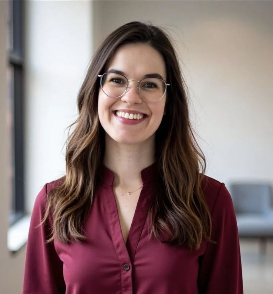A young woman with long wavy brown hair, glasses, and a maroon blouse smiling indoors.