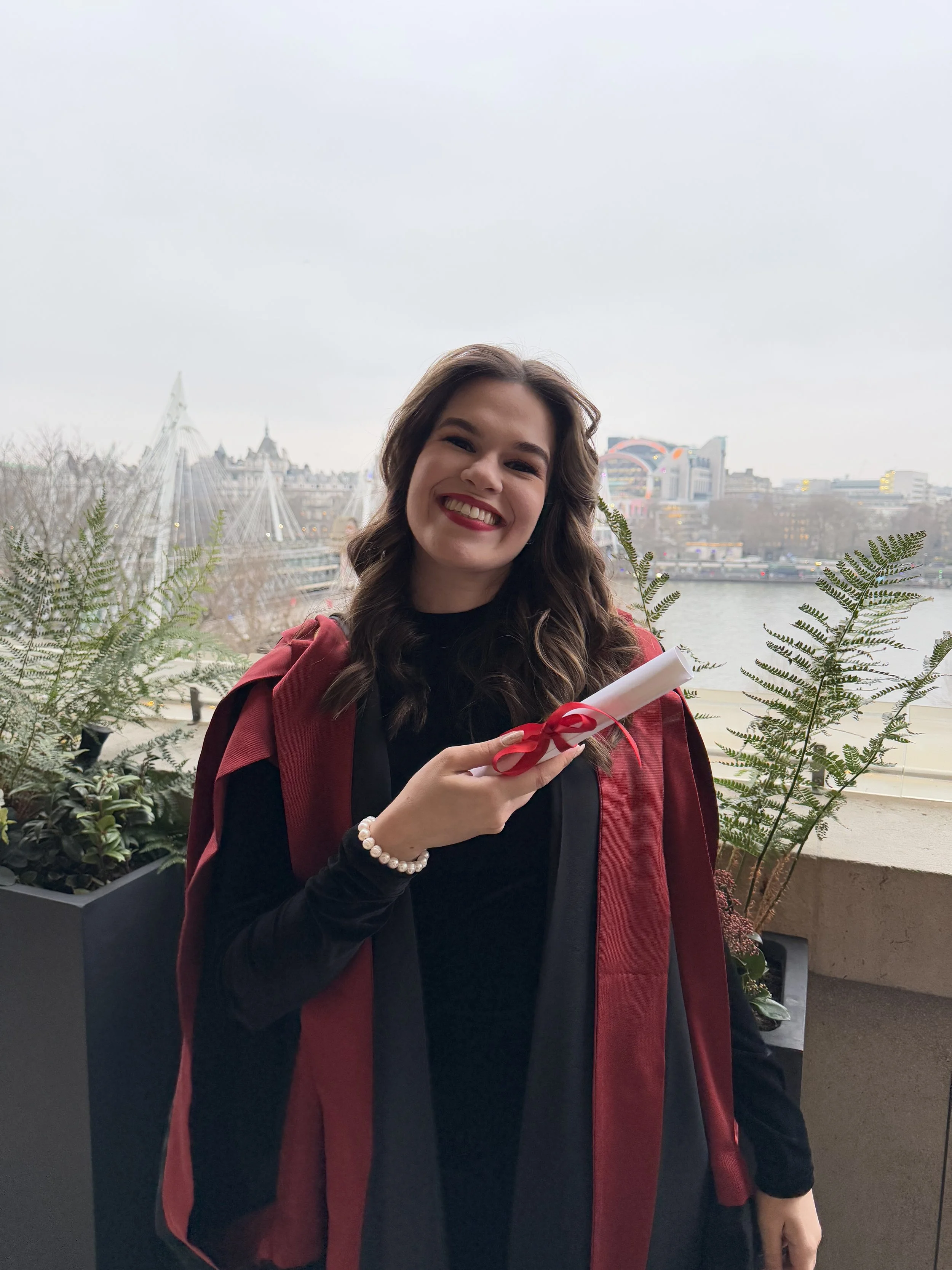 A young woman with brown hair smiling, holding a rolled-up document tied with a red ribbon, in an outdoor setting with a cityscape and a river in the background.