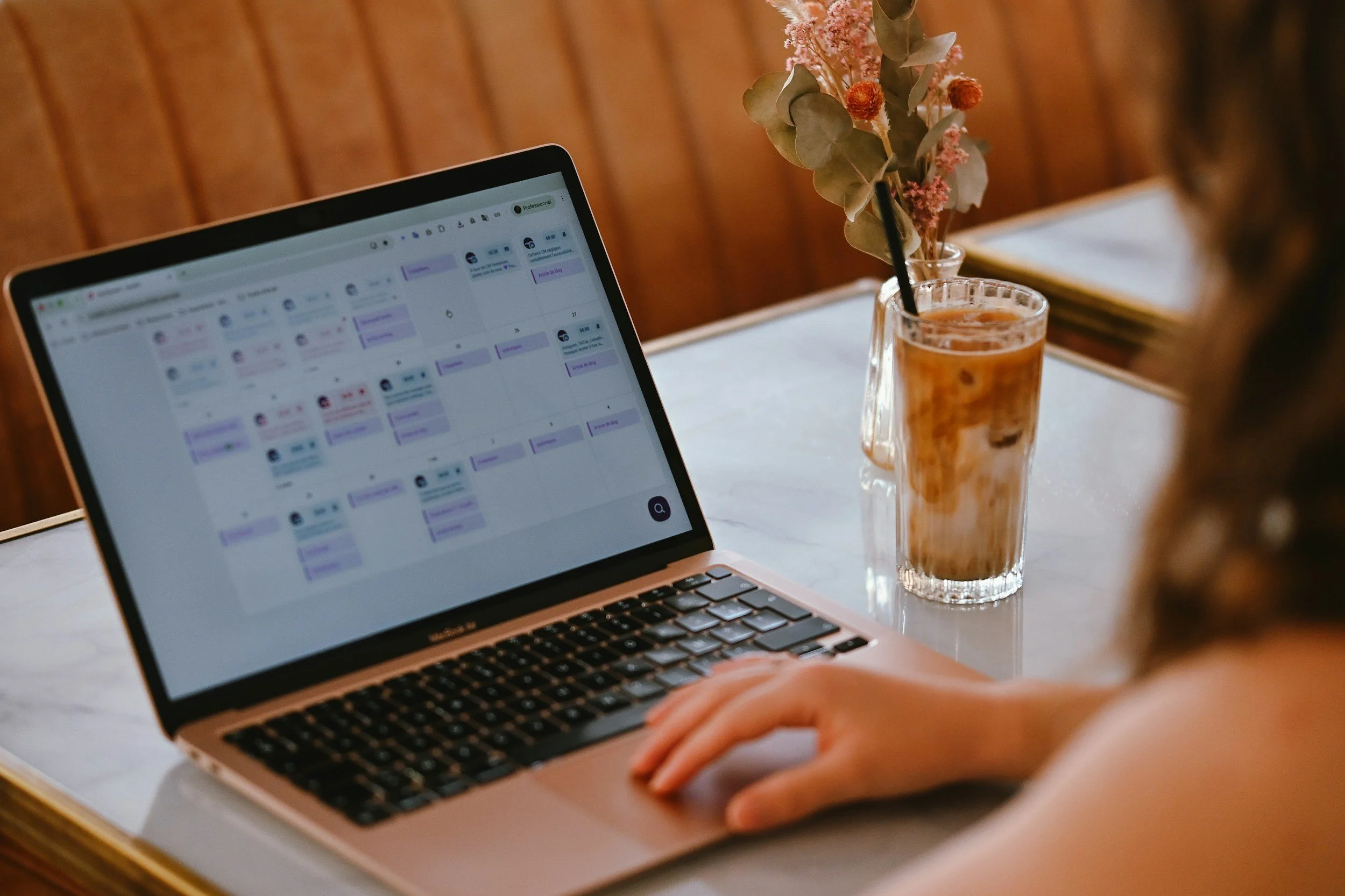 Person working on a laptop displaying a project calendar beside an iced coffee and vase of dried flowers