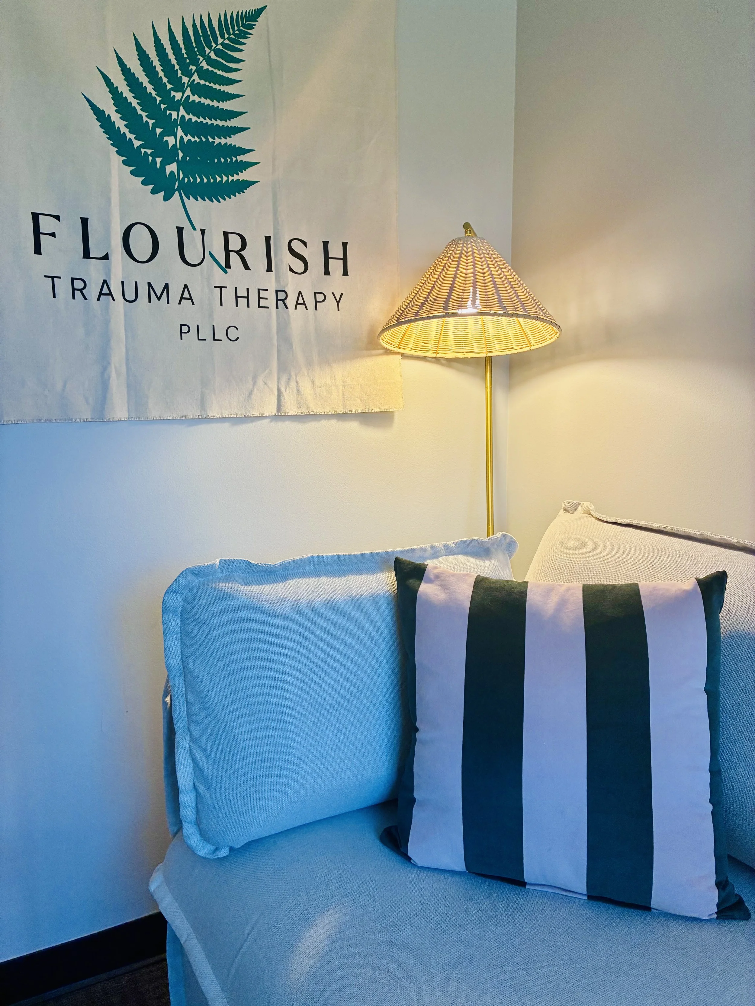 A cozy corner with a couch, a decorative pillow, a wall lamp, and a wall hanging that says 'FLOURISH TRAUMA THERAPY PLLC' with a leaf logo.