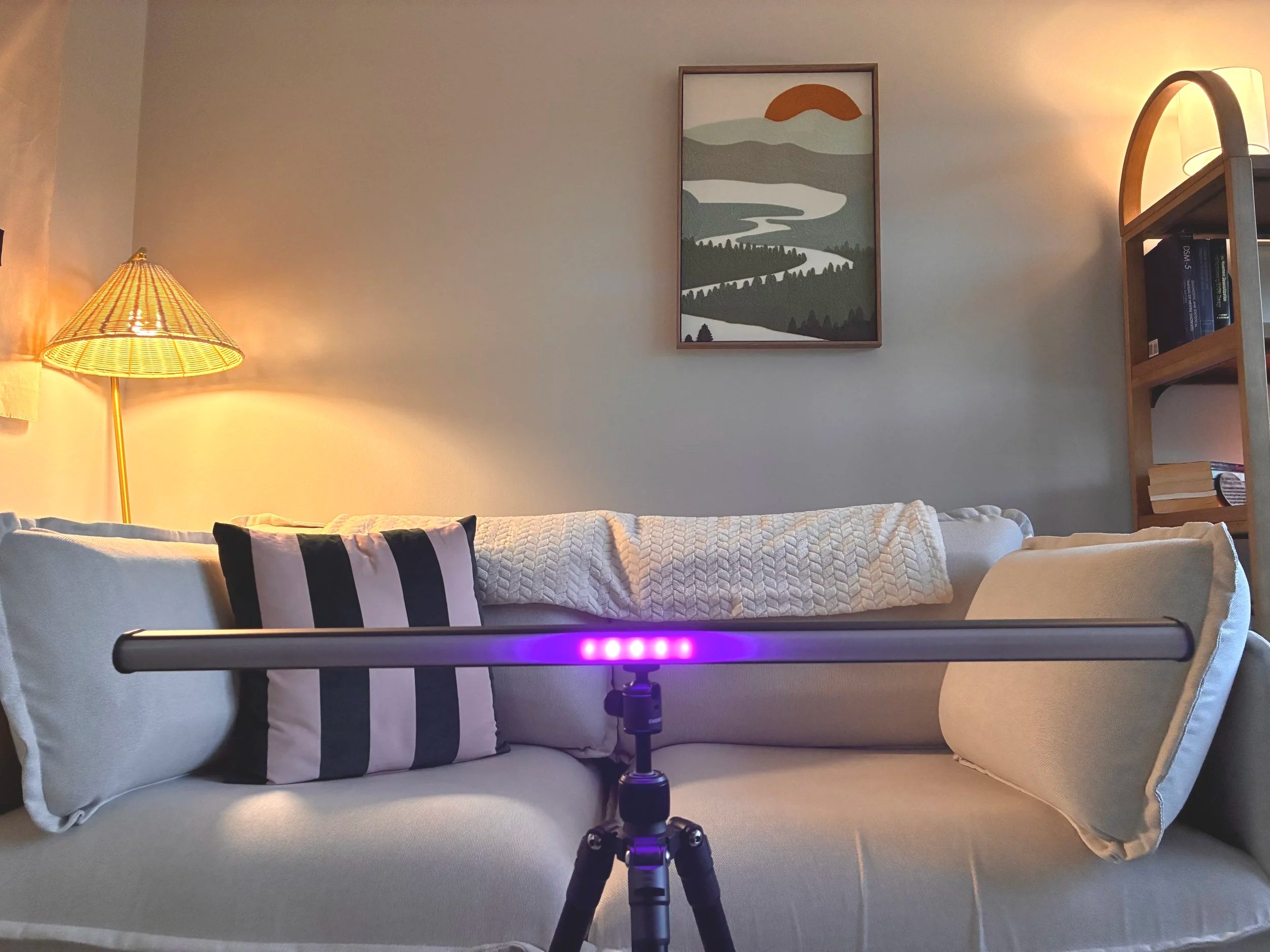 Room with cream-colored sofa, decorative pillows, a floor lamp with a rattan lampshade, framed landscape wall art, and a bookshelf. There is a device with purple lights set up on a tripod in front of the sofa.