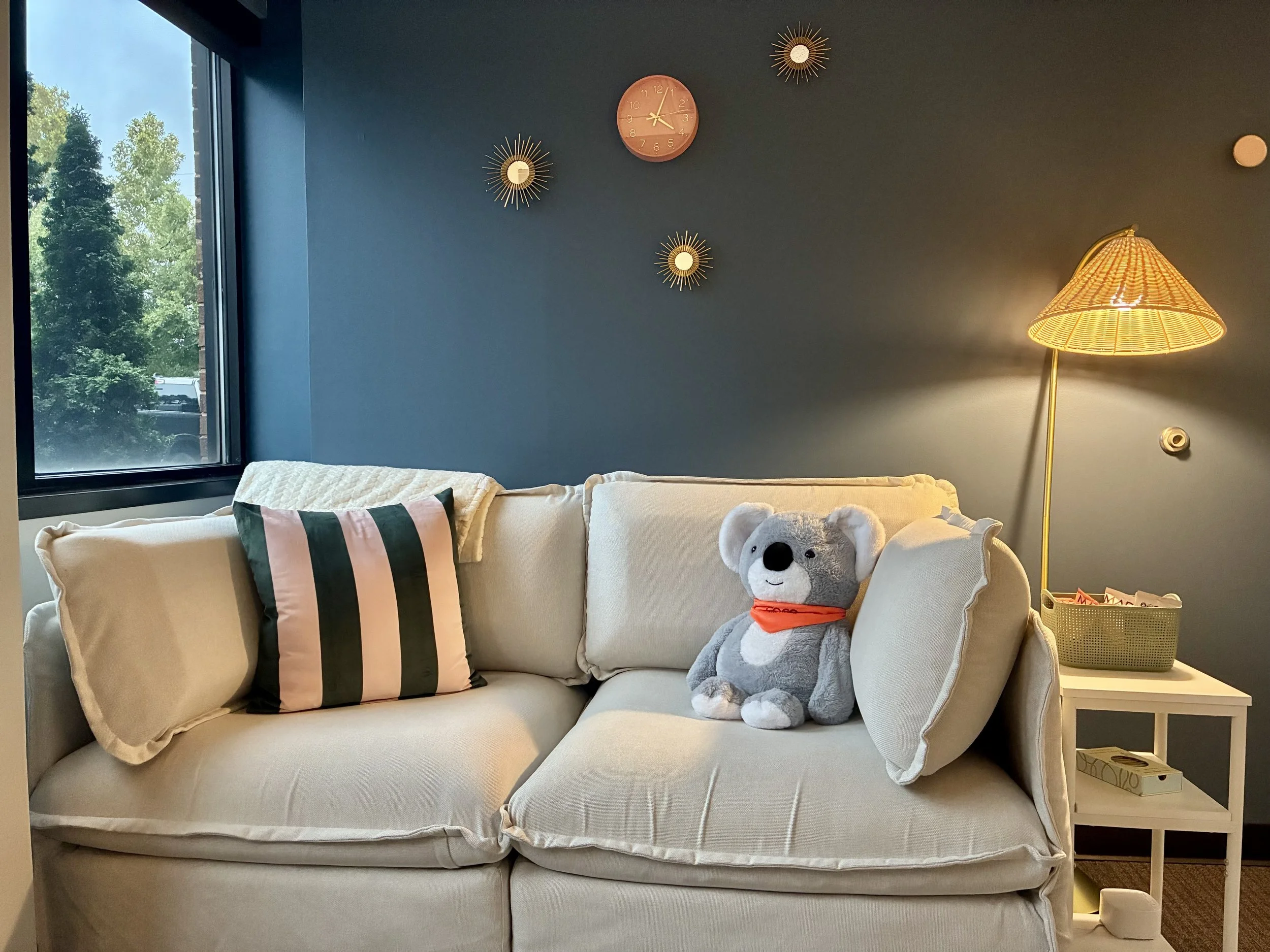 Living room with beige couch, decorative striped pillow, stuffed koala bear with orange bandana, dark blue wall with clock and starburst decorations, window showing trees outside, yellow floor lamp, white side table with a box and decorative items.
