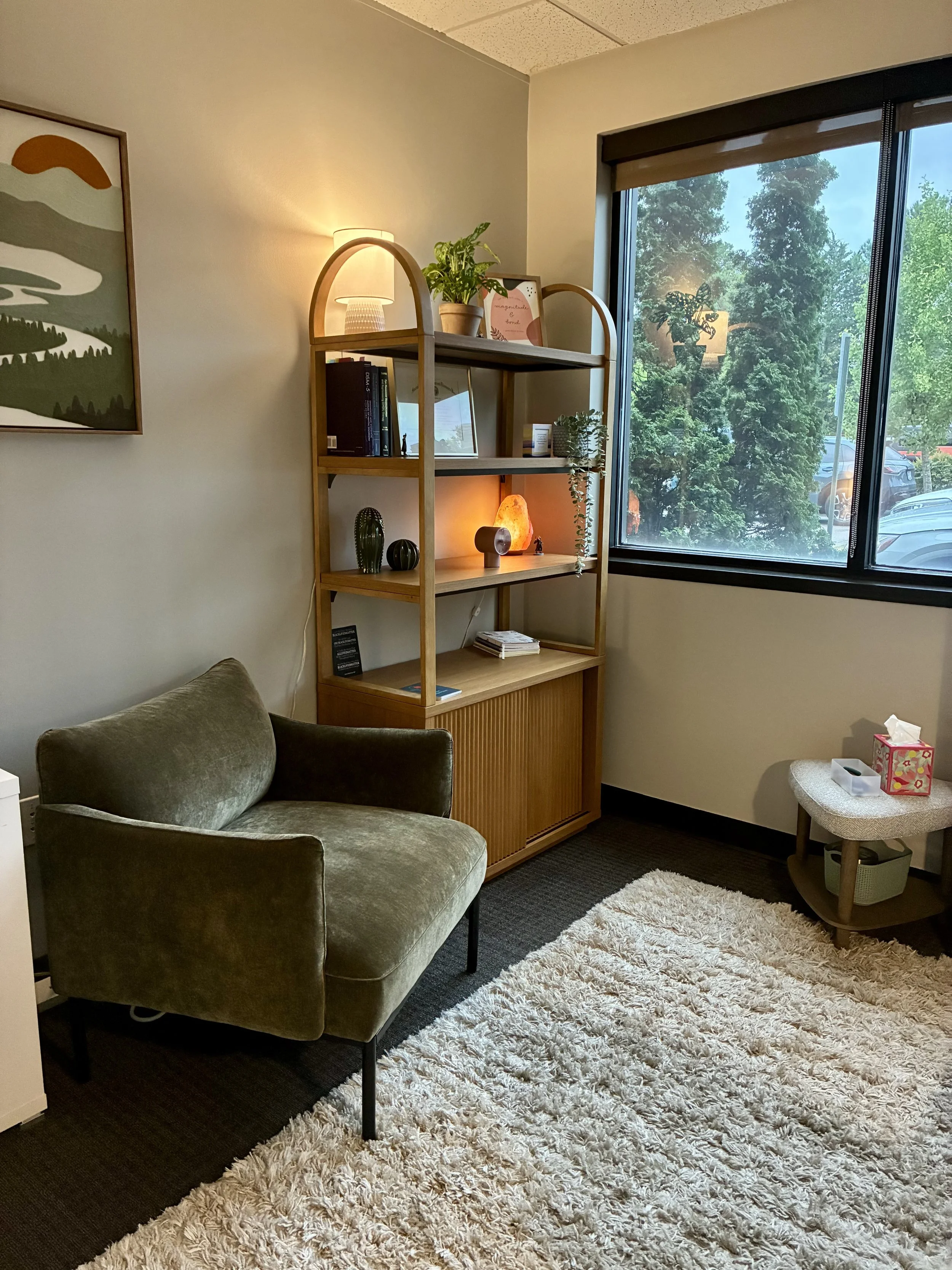 A cozy room corner with a green velvet armchair, a tall wooden shelf with decorative items and plants, a salt lamp, and a large window showing trees outside, with a plush beige rug on the floor.