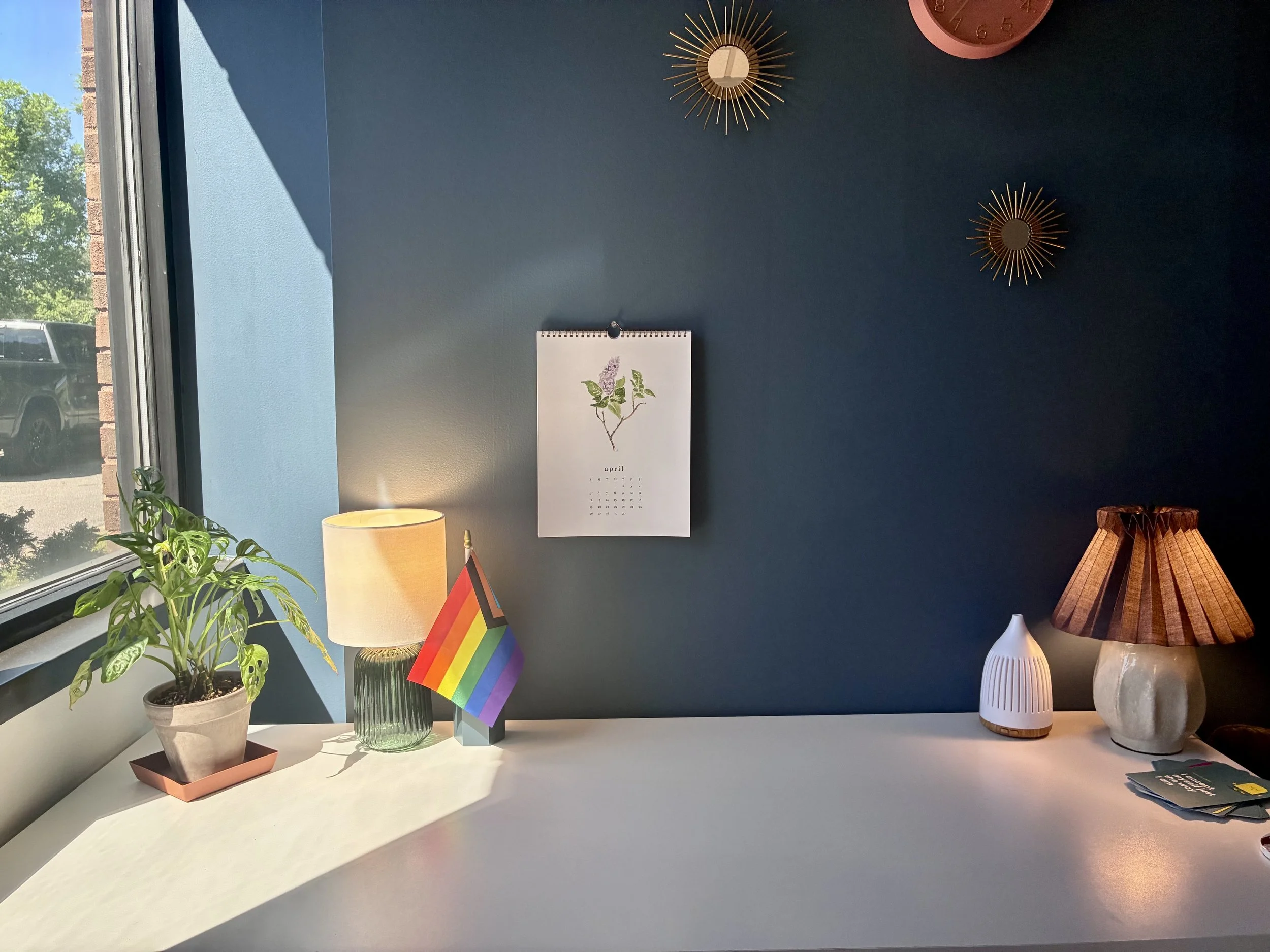 A white table with a potted plant, a glass vase, a small rainbow flag, a lamp, and some books. In the background, a blue wall with three decorative sunburst mirrors and a calendar with the month of April.