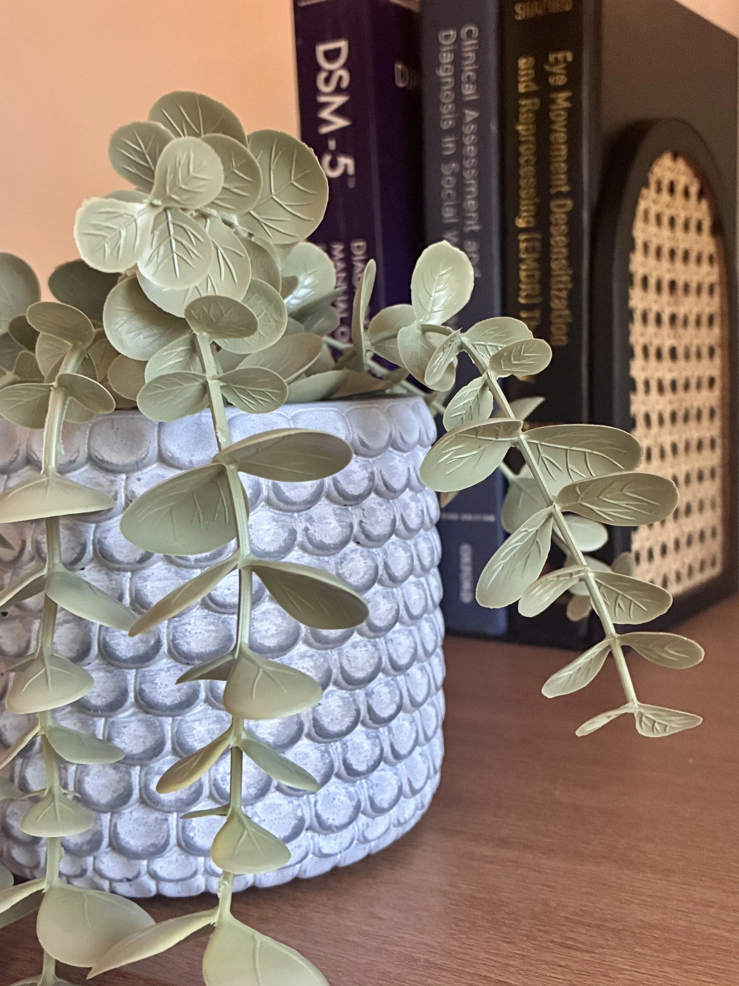 Close-up of a decorative white ceramic pot with faux green leaves, placed on a wooden surface, with books in the background.