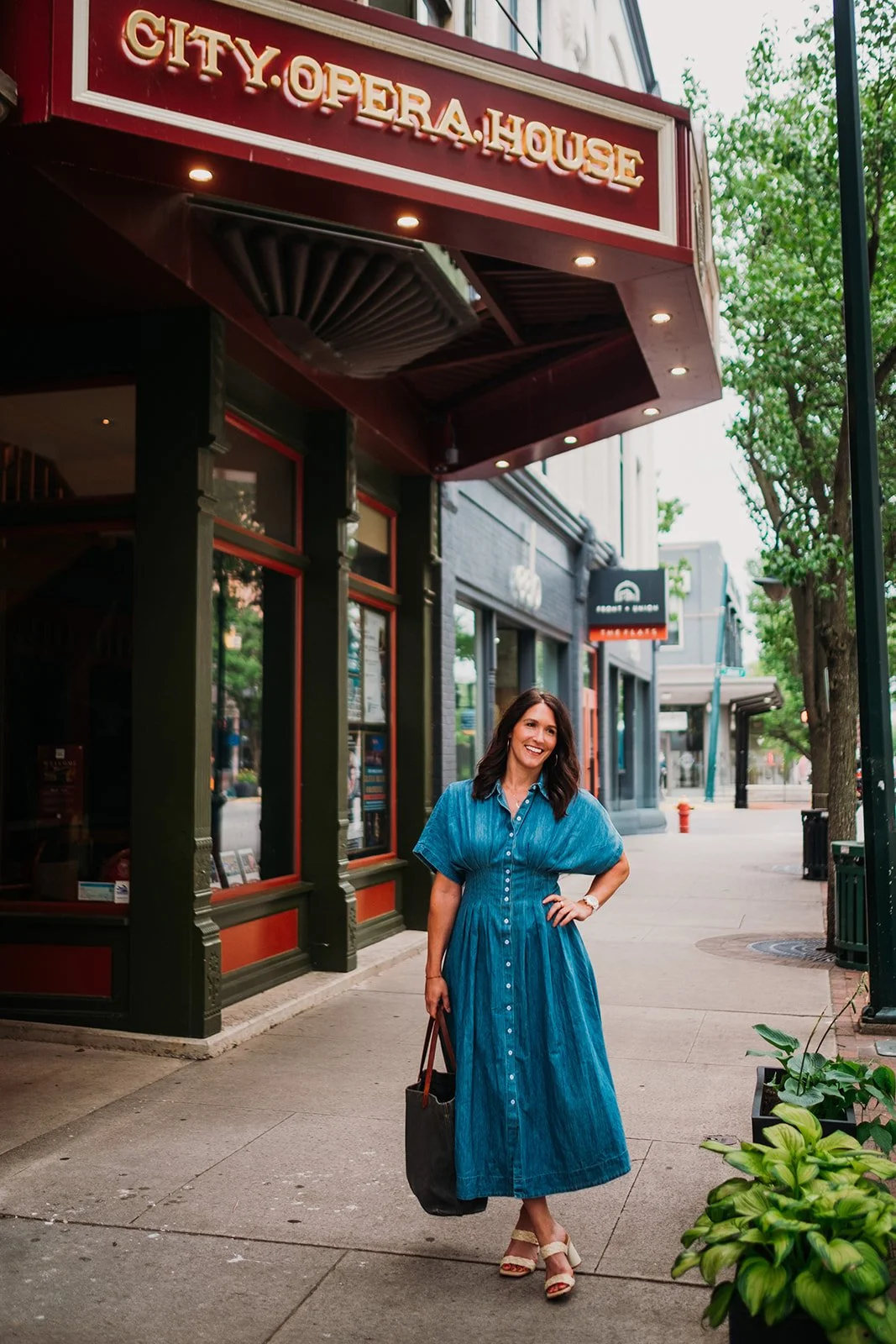 Jenna Morris, TC Wellness Week Founder, in a denim dress standing on the sidewalk outside a theatre entrance with a sign reading 'City Opera House'.