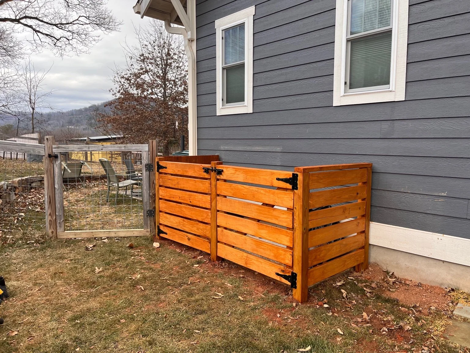 A small wooden fence with a gate attached to the side of a gray house with white trim. The backyard has uneven grass and dried leaves, surrounded by a wire fence with wooden posts. There are chairs and trees in the background, with hills and an overcast sky.