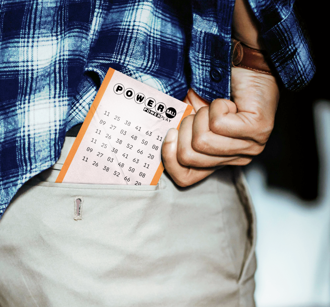 Close-up of a person wearing a blue plaid shirt and beige pants, holding a lotto ticket in their front pocket.