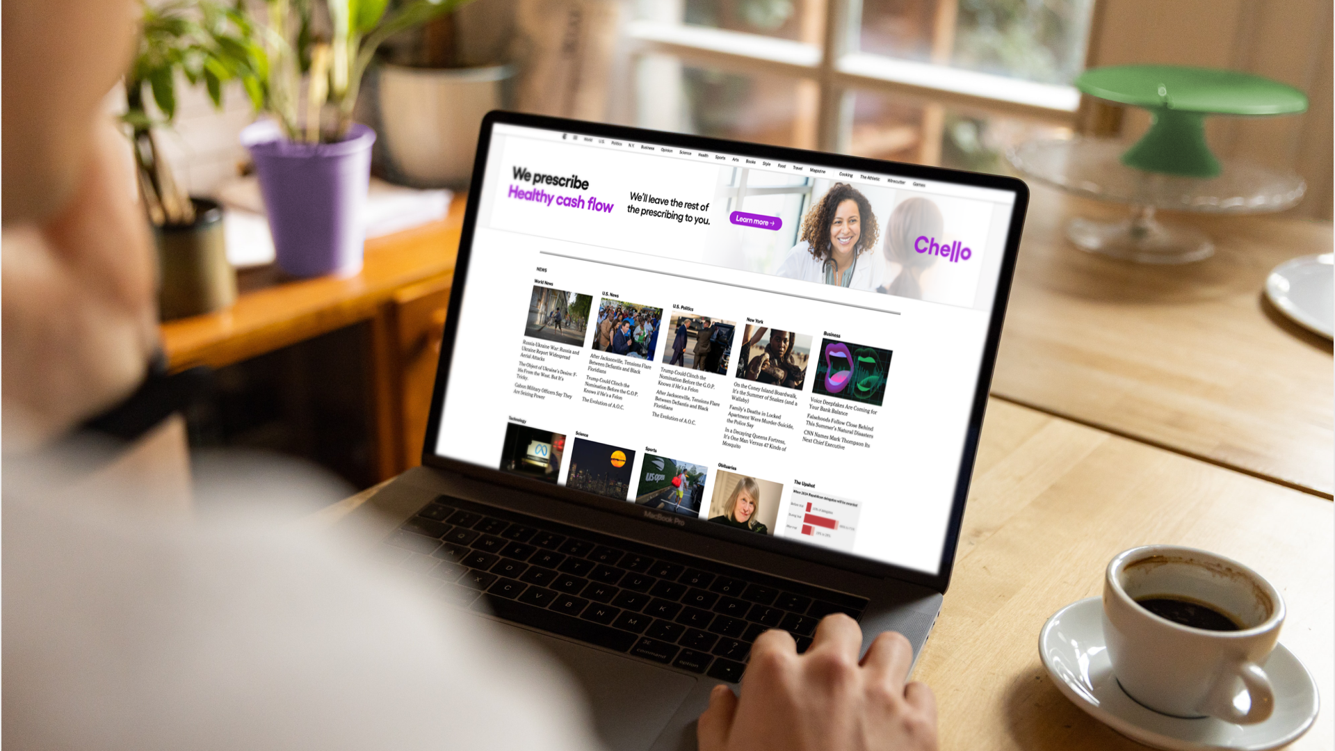 A person using a laptop on a wooden table shows a news website. The website features news headlines and images, with a header advertisement stating "We prescribe Healthy cash flow" and a purple "Learn more" button. A cup of coffee is on a saucer to the right, and a green desk lamp and potted plants are visible in the background.