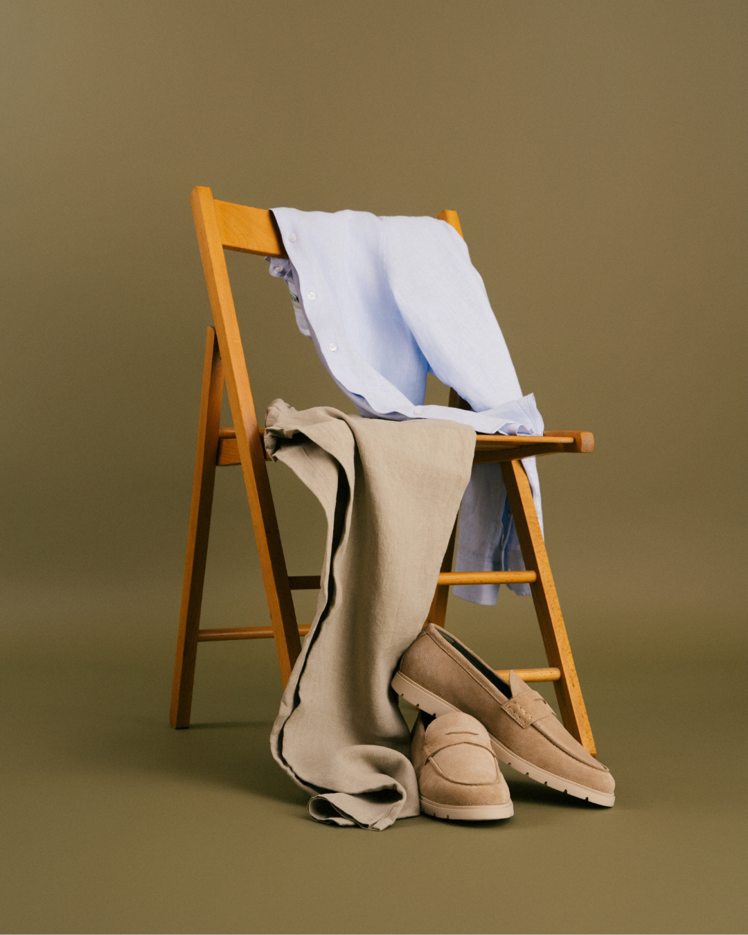 Clothing and shoes draped over a wooden folding chair against a green background, including a light blue shirt, beige pants, and tan loafers.