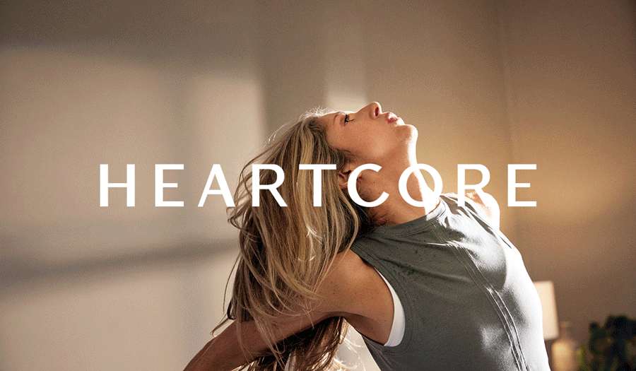 A woman with long blonde hair stretching, facing upward with eyes closed in a warmly lit room with the word 'HEARTCORE' overlaid.