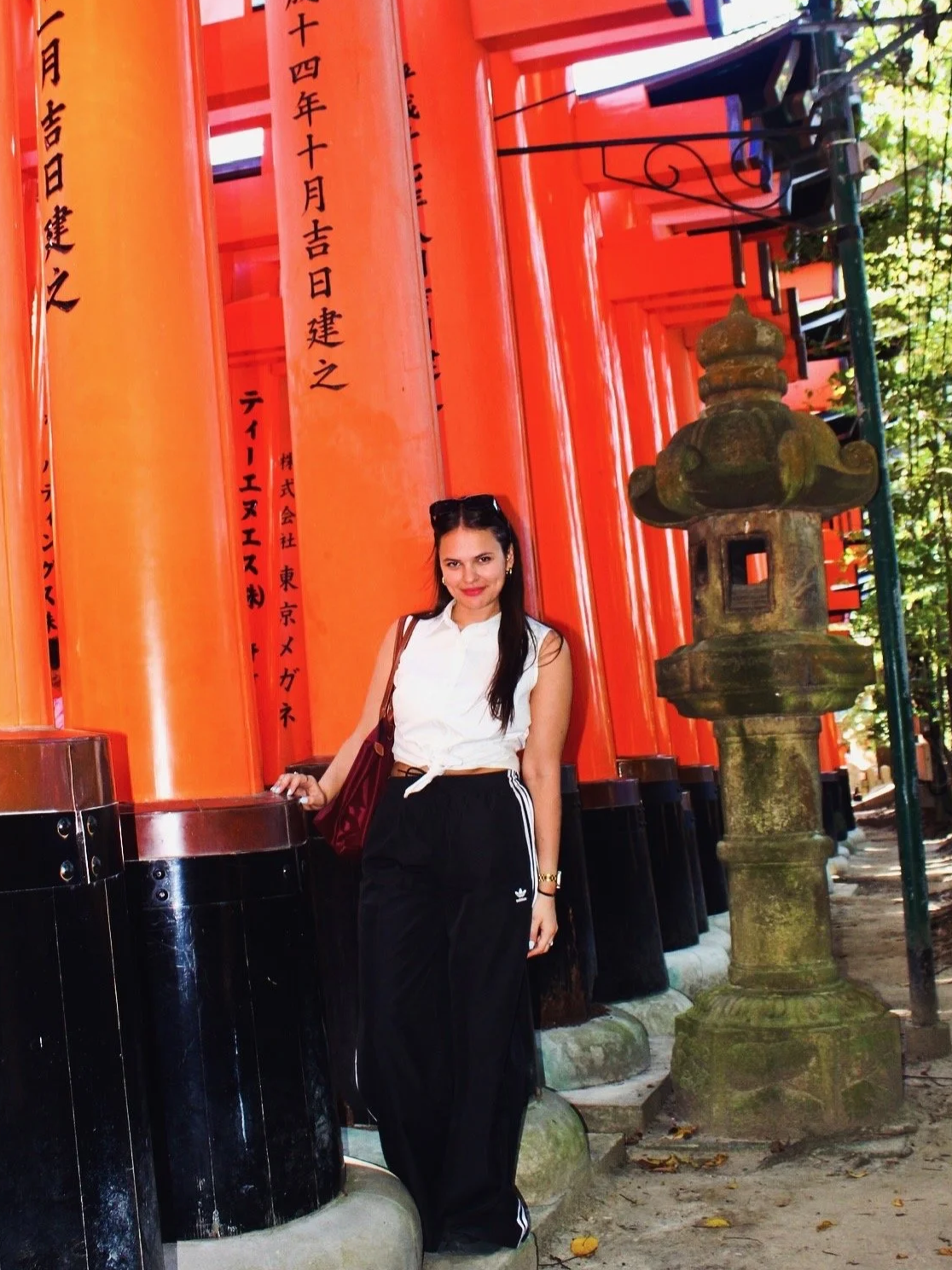 Woman standing in front of a row of bright orange torii gates at a Japanese shrine, holding onto one of the gates with her hand.