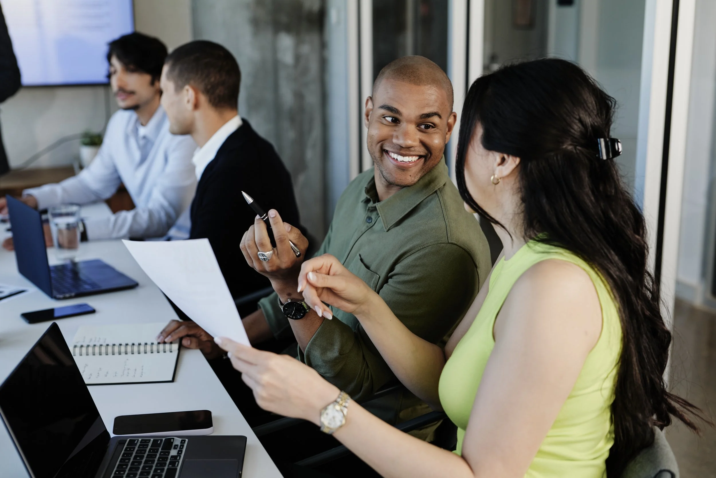 Two colleagues, a man and a woman, are engaged in conversation during a work meeting in an office conference room. The man is smiling and holding a pen, while the woman is gesturing and holding a paper. Other colleagues are seated in the background, working on laptops.