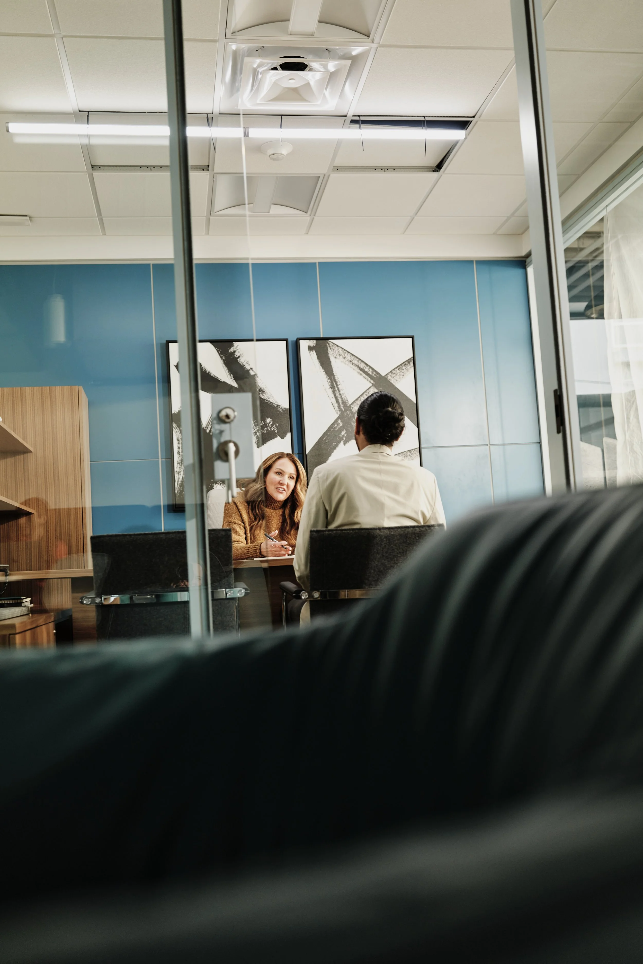 A woman and a man sitting at a table in an office, engaged in a conversation. The woman is holding a pen, and the background features a blue wall with abstract black and white art.