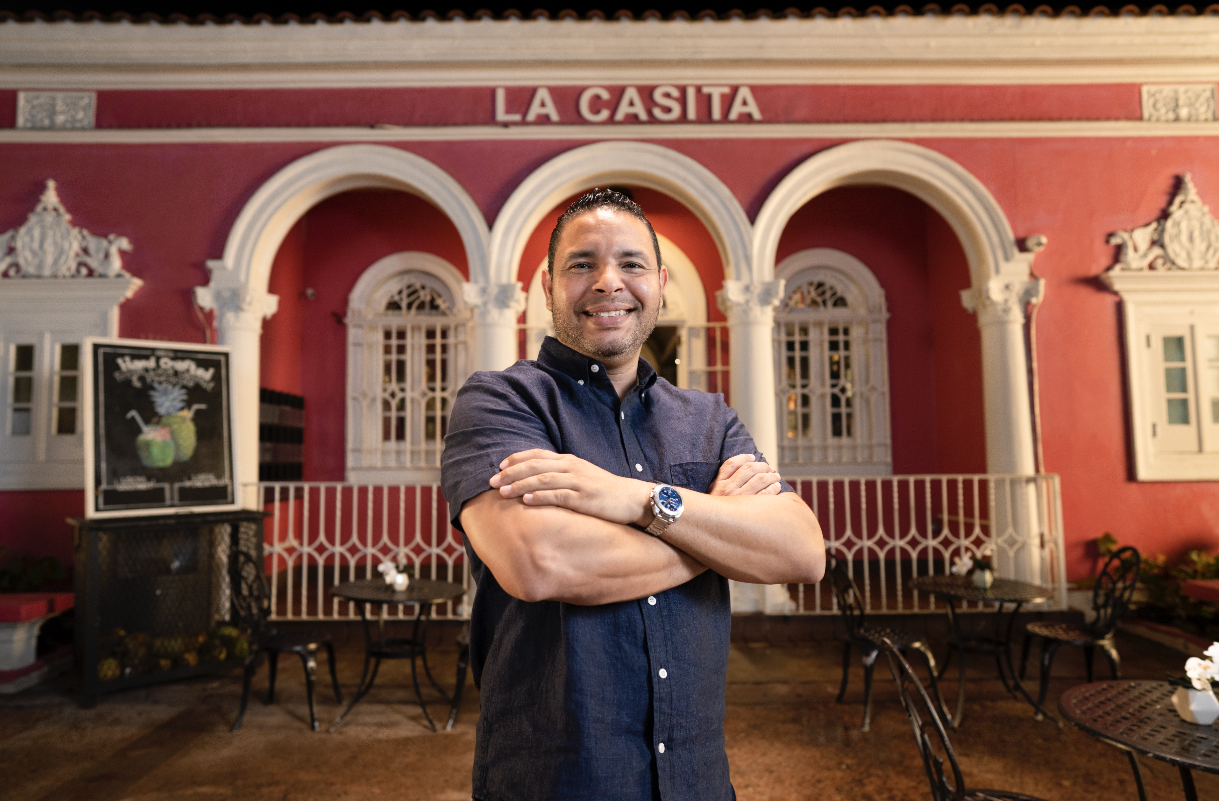 A smiling man with arms crossed, wearing a wristwatch and a dark button-up shirt, standing in front of a red building with white decorative architecture and the sign 'LA CASITA'