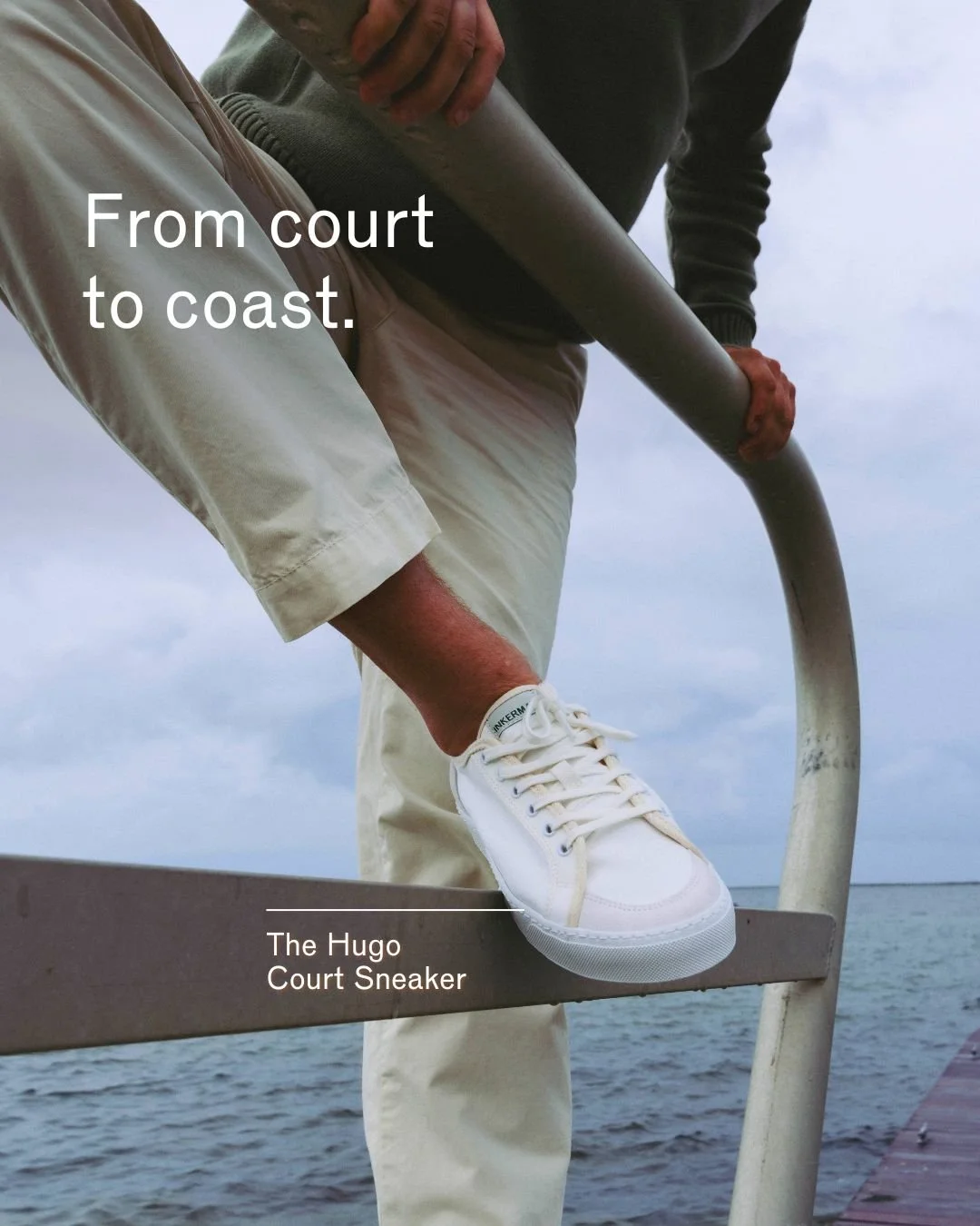 Close-up of a person wearing white sneakers and beige pants, standing on a dock with water in the background, while another person holding a pole stands nearby. Text on the image reads 'From court to coast.' and 'The Hugo Court Sneaker'.
