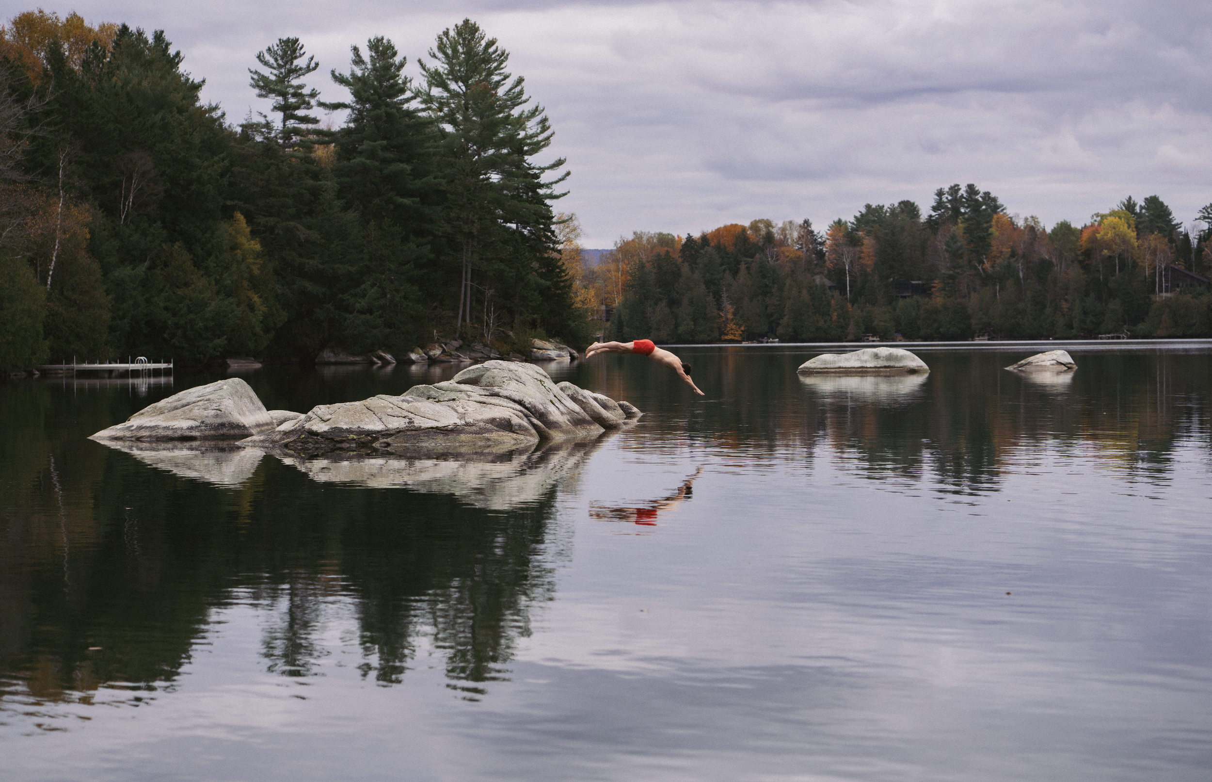 A person diving into a lake with rocks and trees in the background, overcast sky.