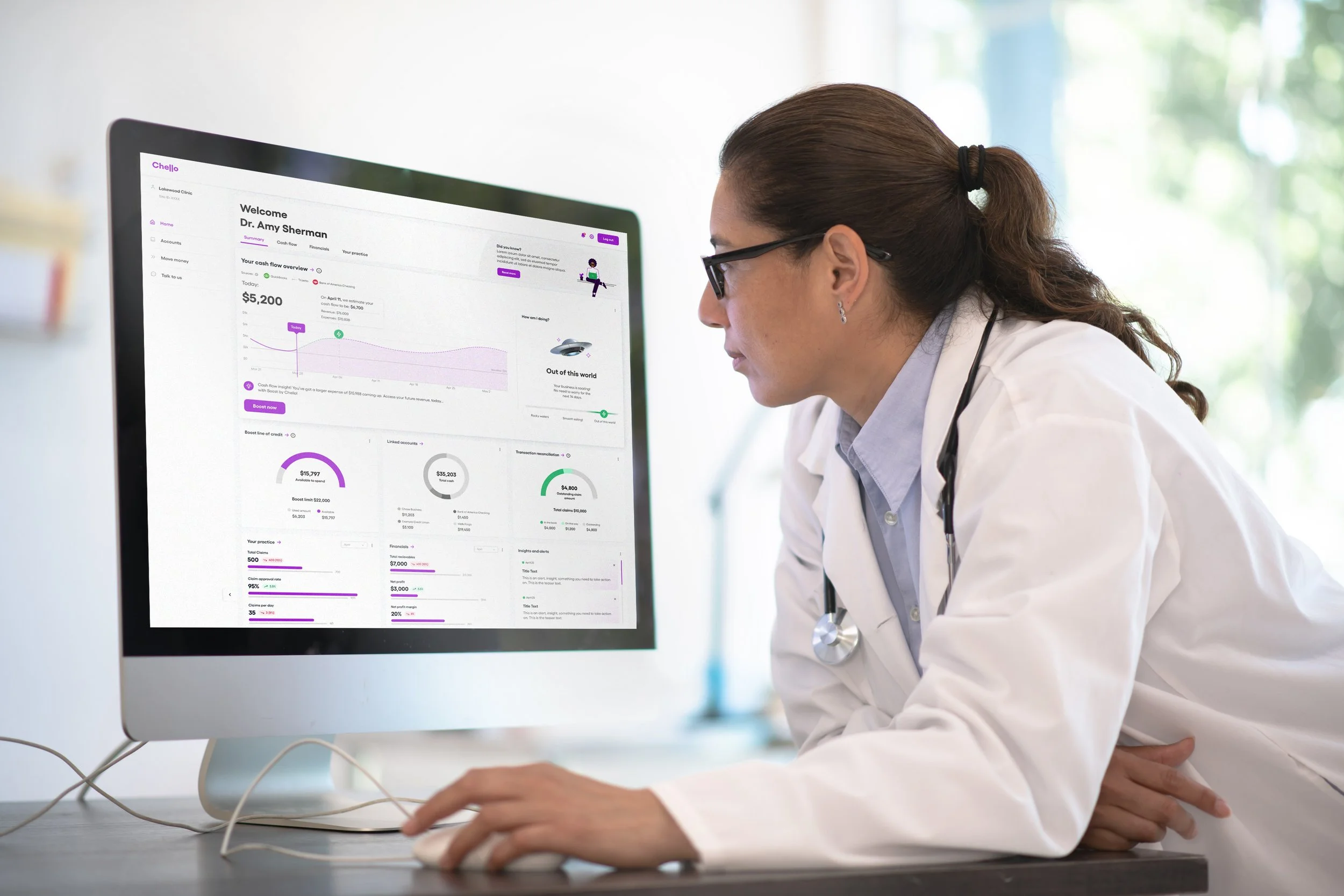 A female doctor wearing a white coat and glasses is looking at a computer monitor displaying an online dashboard or medical data.