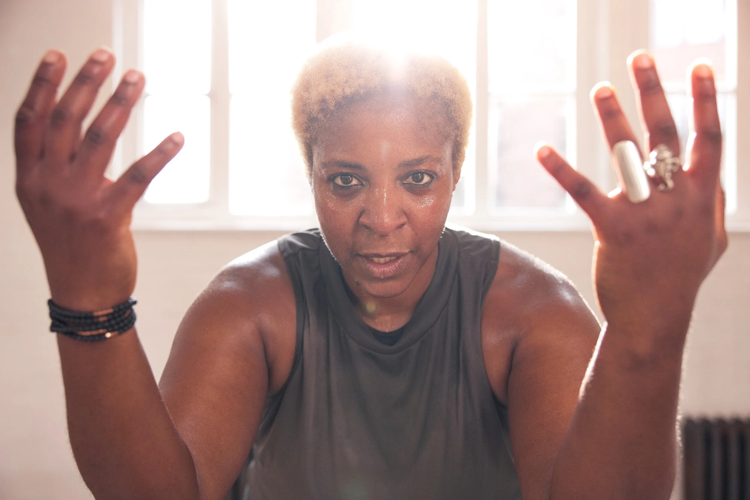 A woman with short blonde hair and dark skin, sweating and looking directly at the camera, with her hands raised.