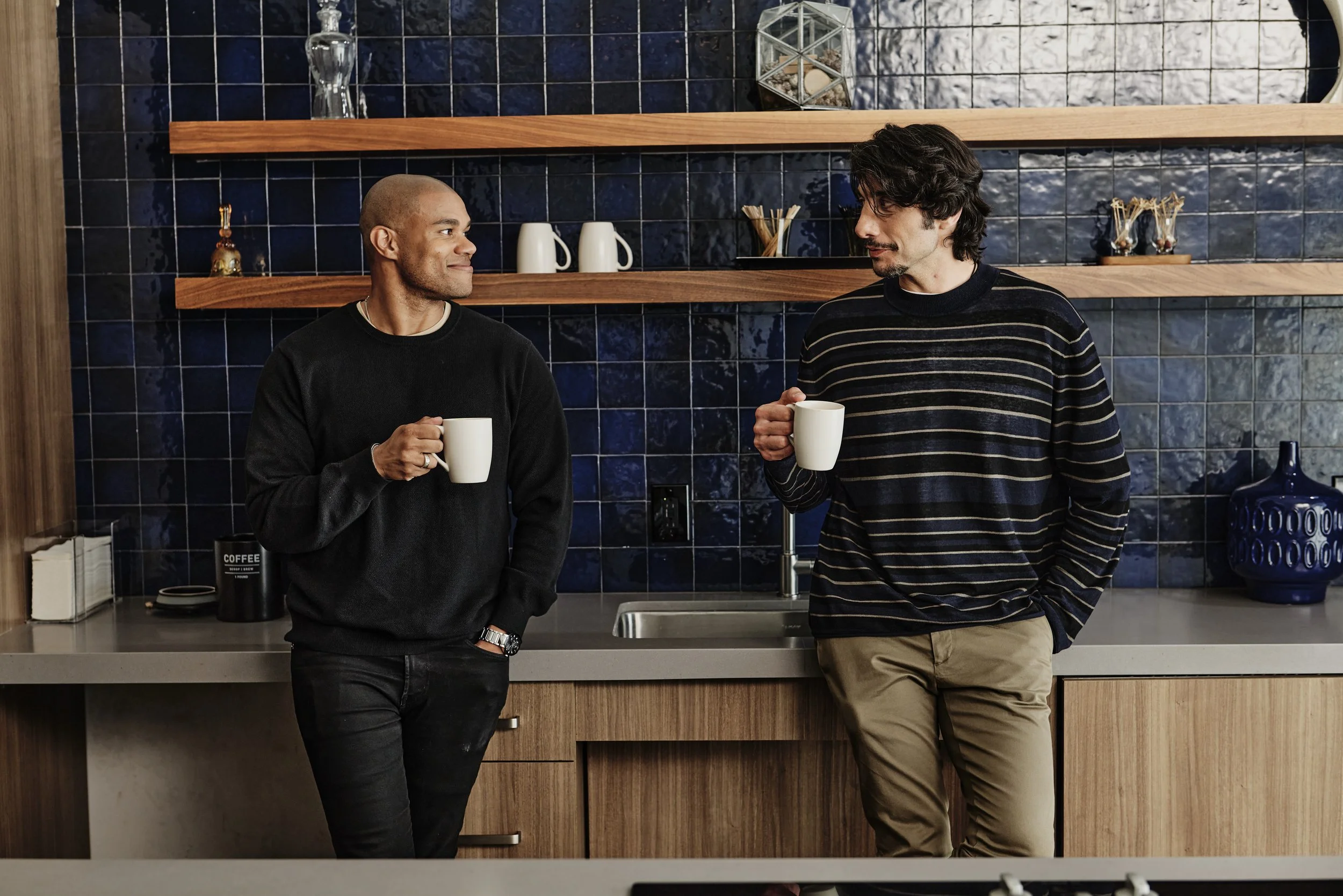 Two men are standing in a kitchen, holding white coffee mugs and talking. The man on the left has a shaved head and is wearing a black sweater, while the man on the right has dark wavy hair and is wearing a dark striped sweater. The kitchen features dark blue tiled backsplash, open wooden shelves with decorative items, and a beige countertop.